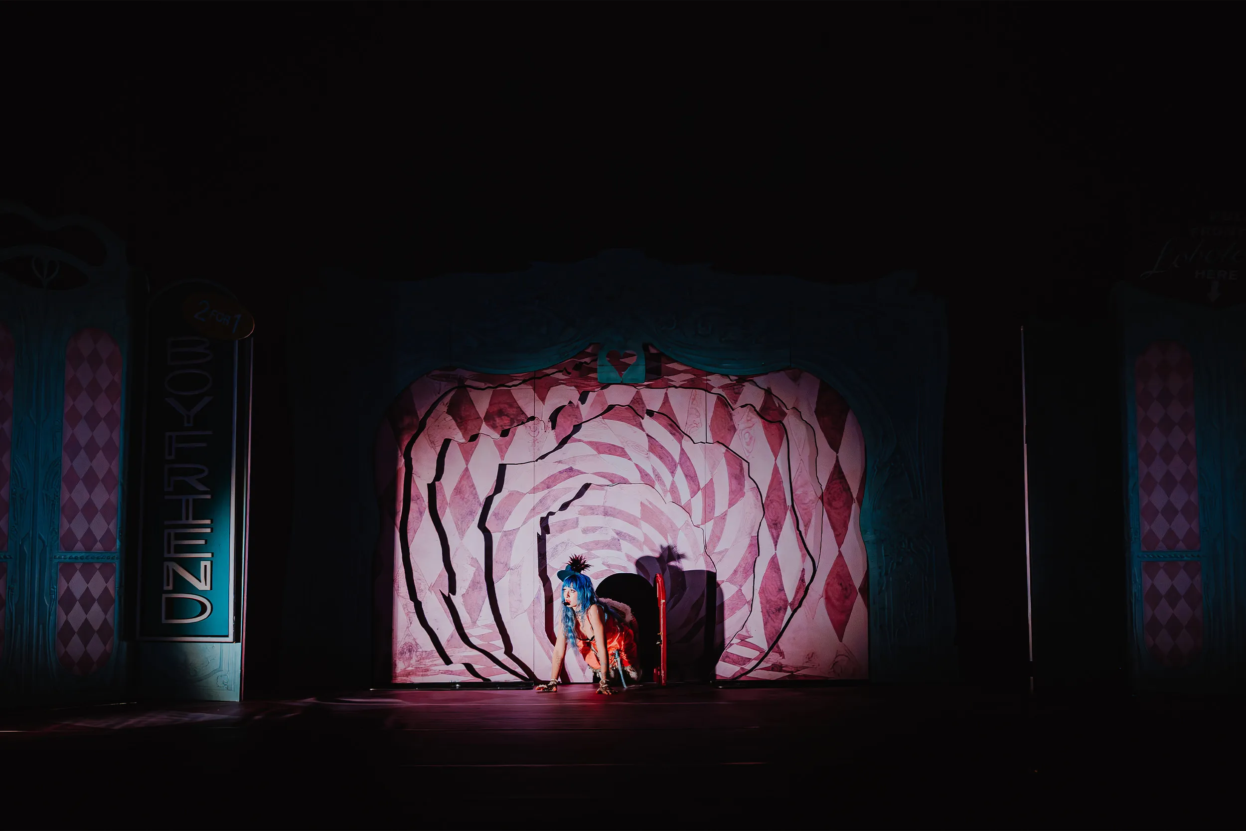 Ashnikko crawling out of a small doorway at the centre of a pink harlequin rose, the rest of the stage in darkness