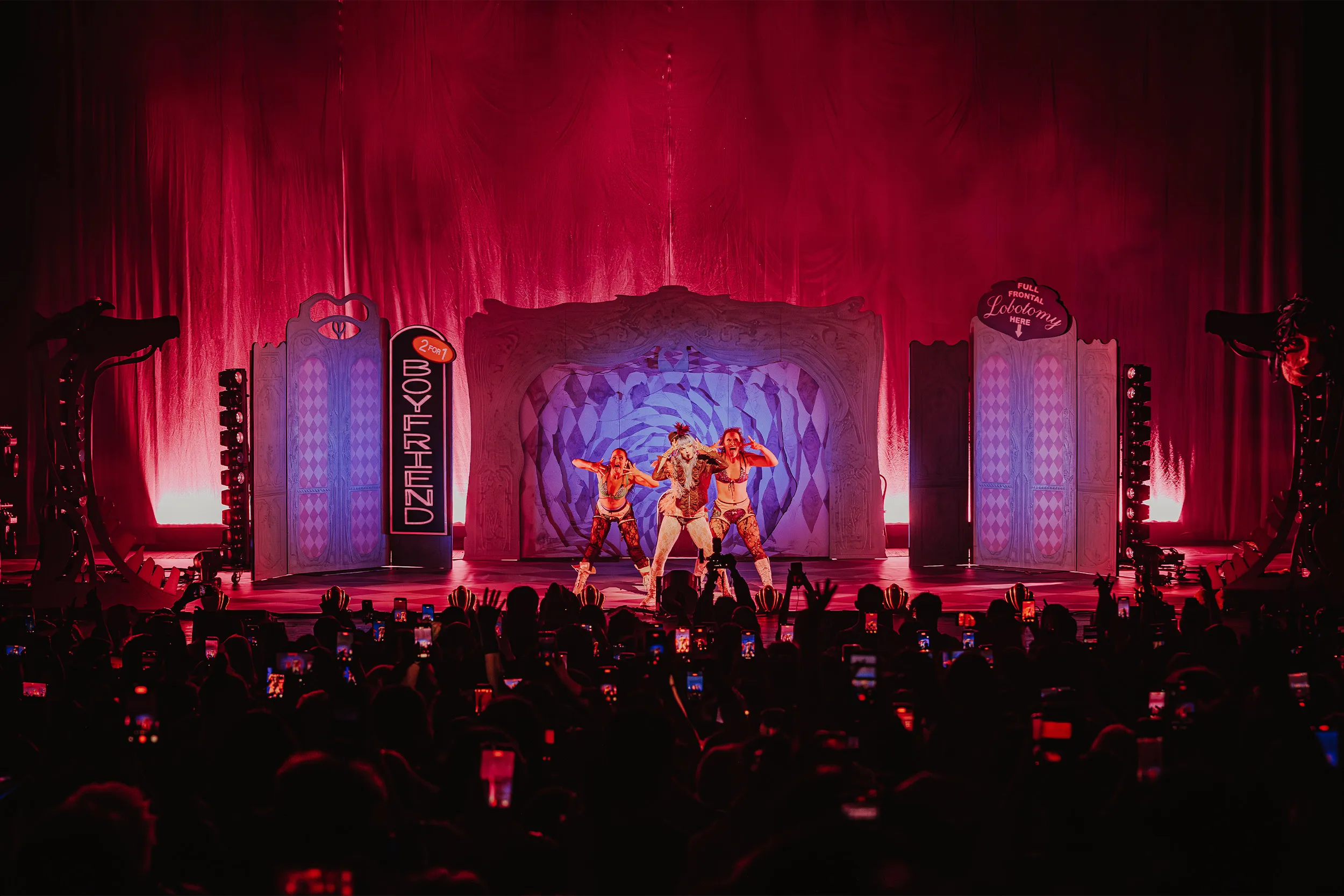Wide shot of the full set under red light: the Boyfriend sign, carnival flats, a lit harlequin rose at centre and a crowd of raised phones