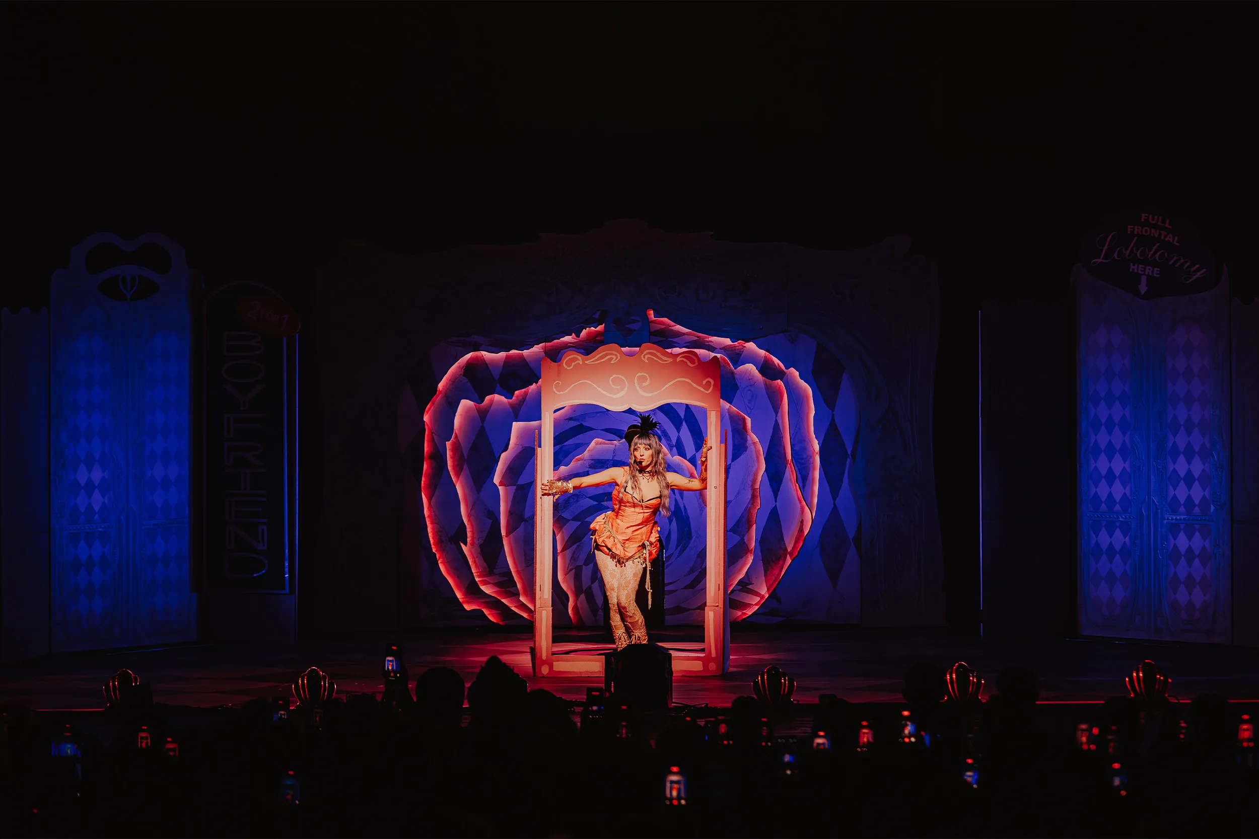 Ashnikko standing in a pink mirror frame lit orange, a blue and pink harlequin rose behind