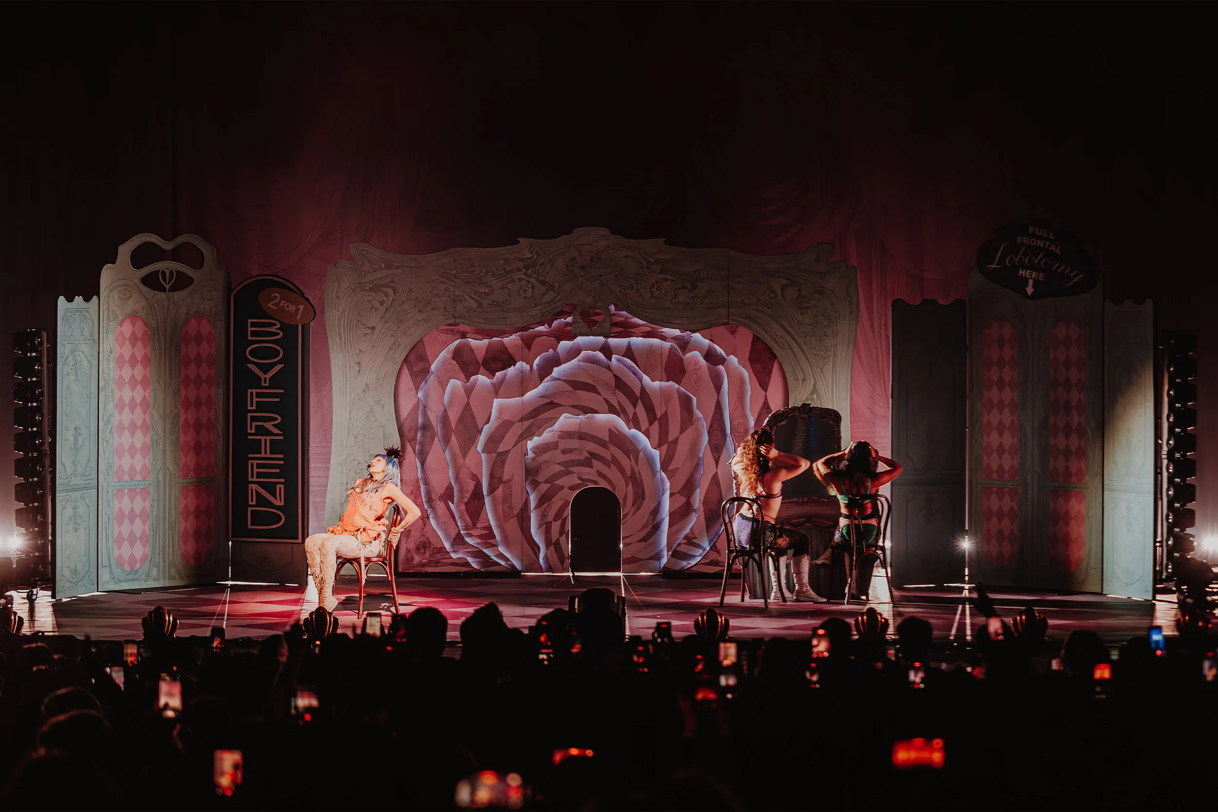 Ashnikko seated on a chair stage left while two dancers sit astride chairs opposite, the rose glowing pink between them