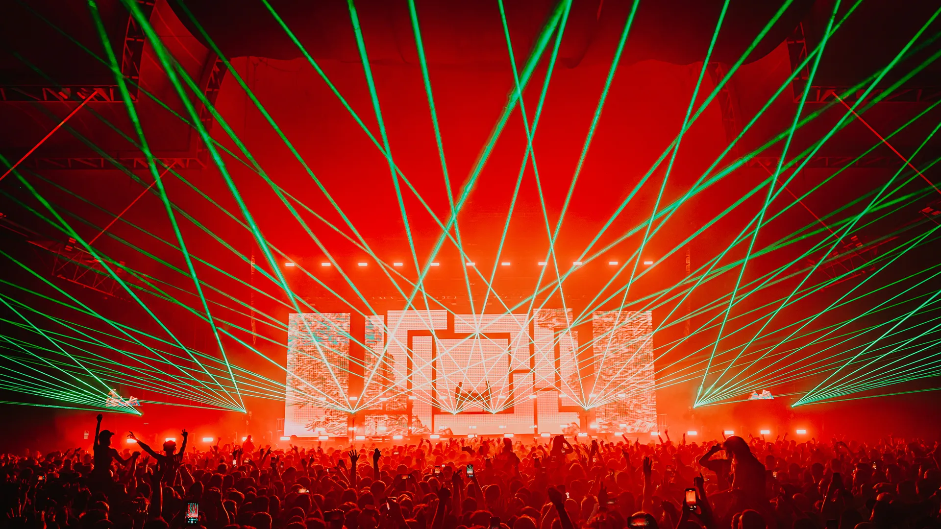 A packed arena in red light with green lasers fanning over the crowd