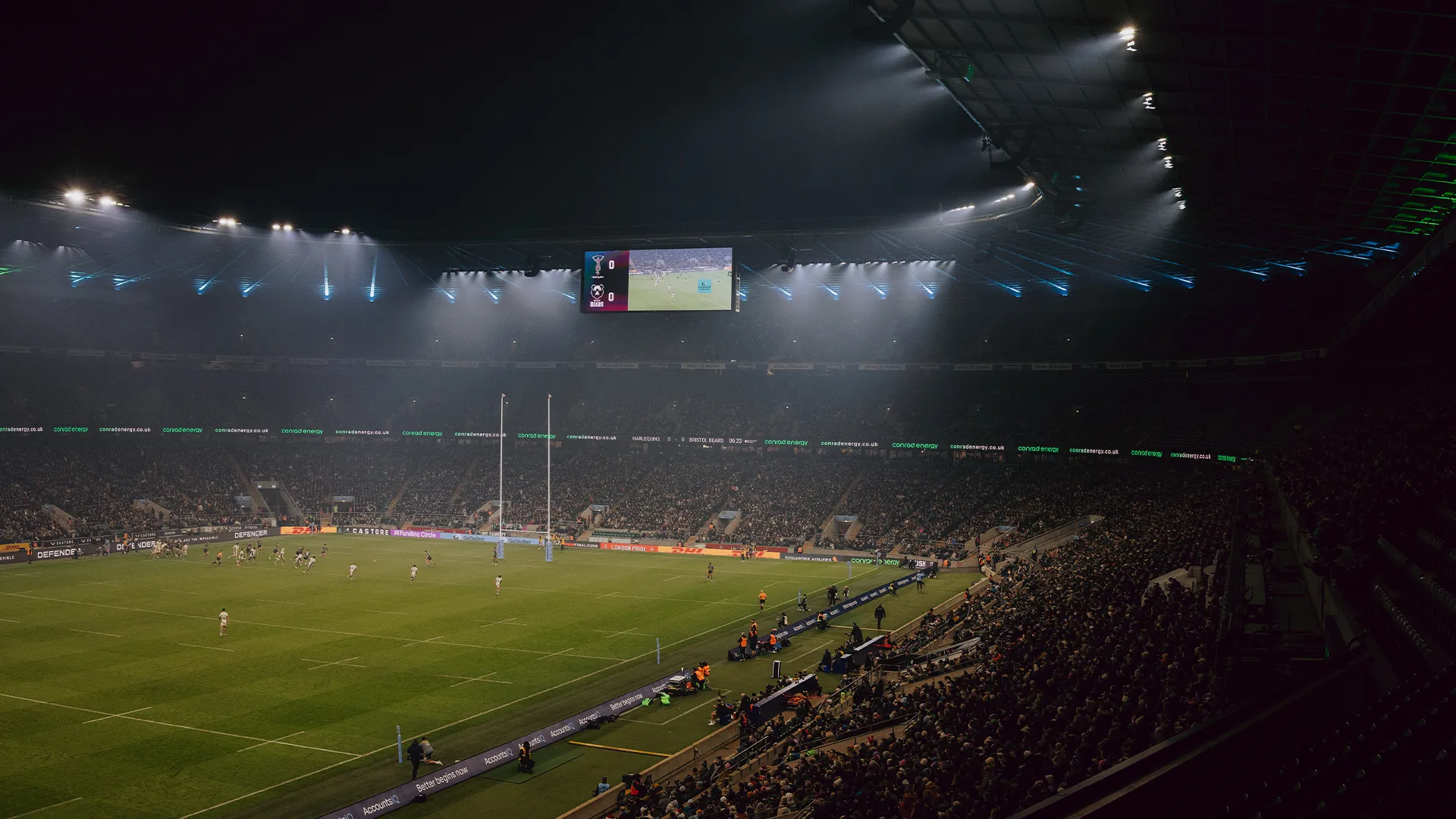 Floodlit rugby match at night, big screen showing the action