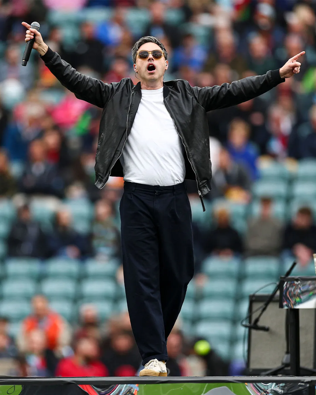 A singer in sunglasses and a leather jacket, arms flung wide, performing in front of packed stands