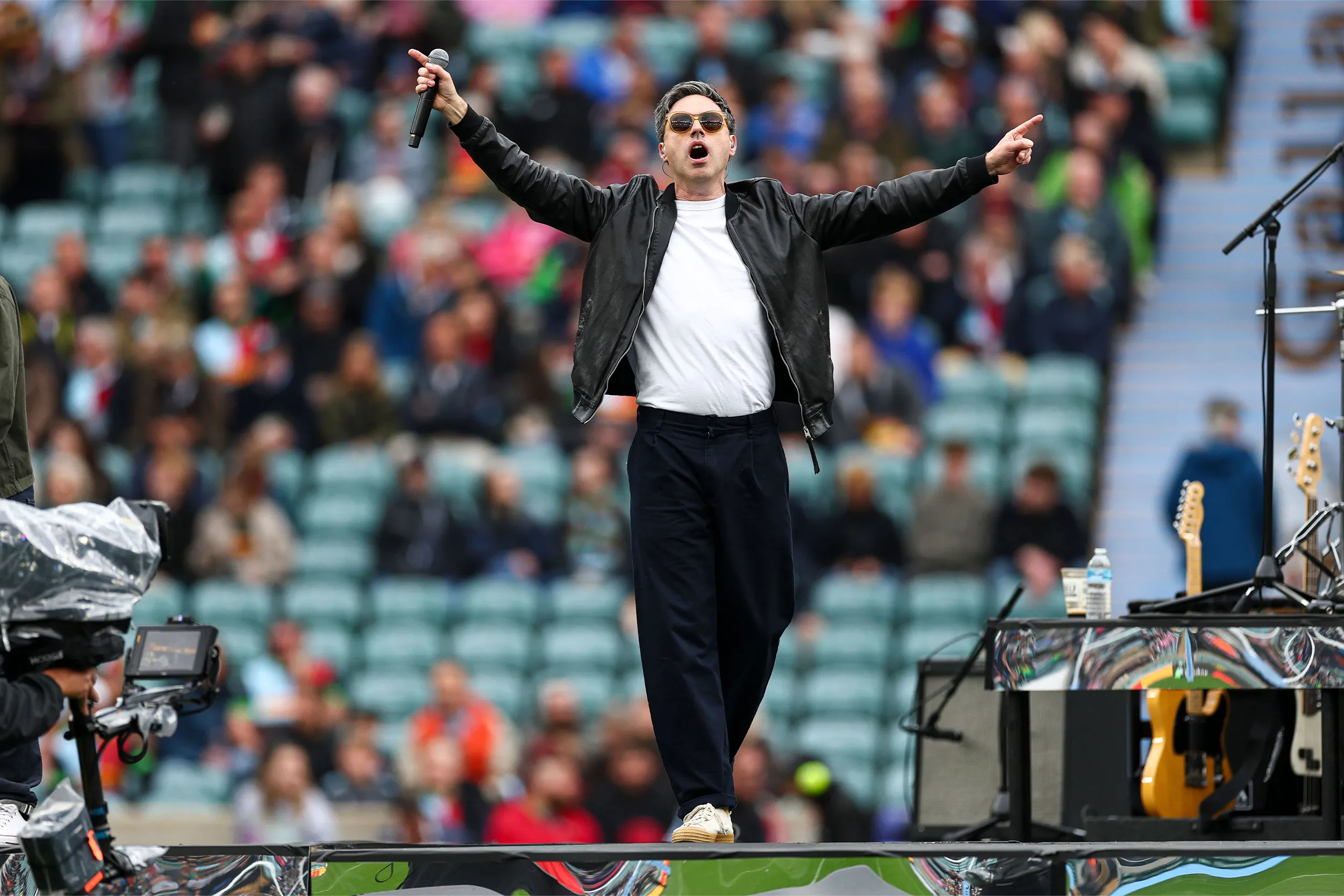 A singer in sunglasses and a leather jacket, arms wide, on a stage in front of the stands