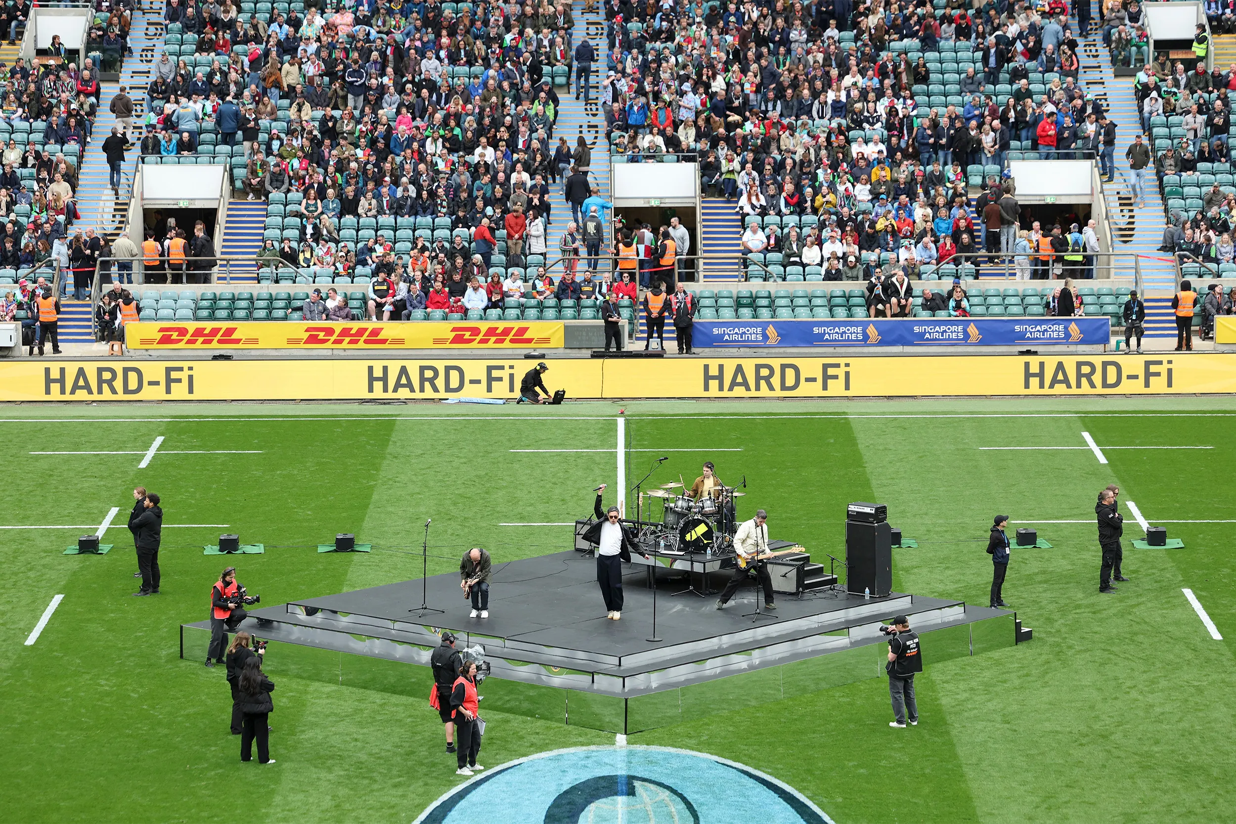 A band playing on a low stage set out on the pitch, camera crew around it and the stands behind