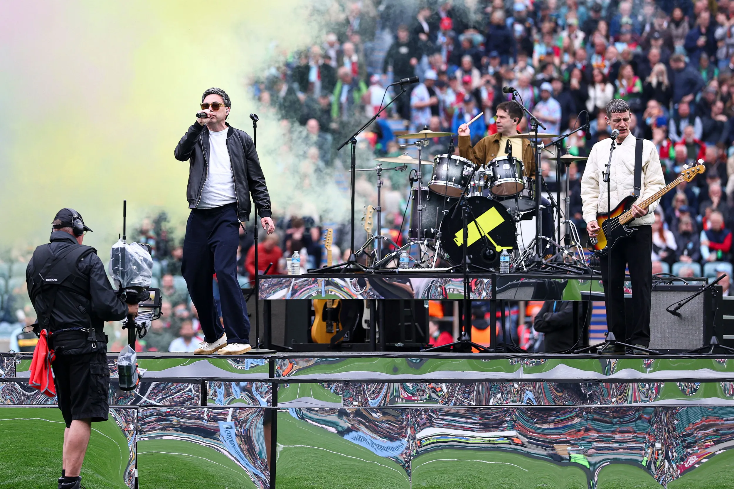The band on the pitch stage in coloured smoke, a camera operator filming from the side