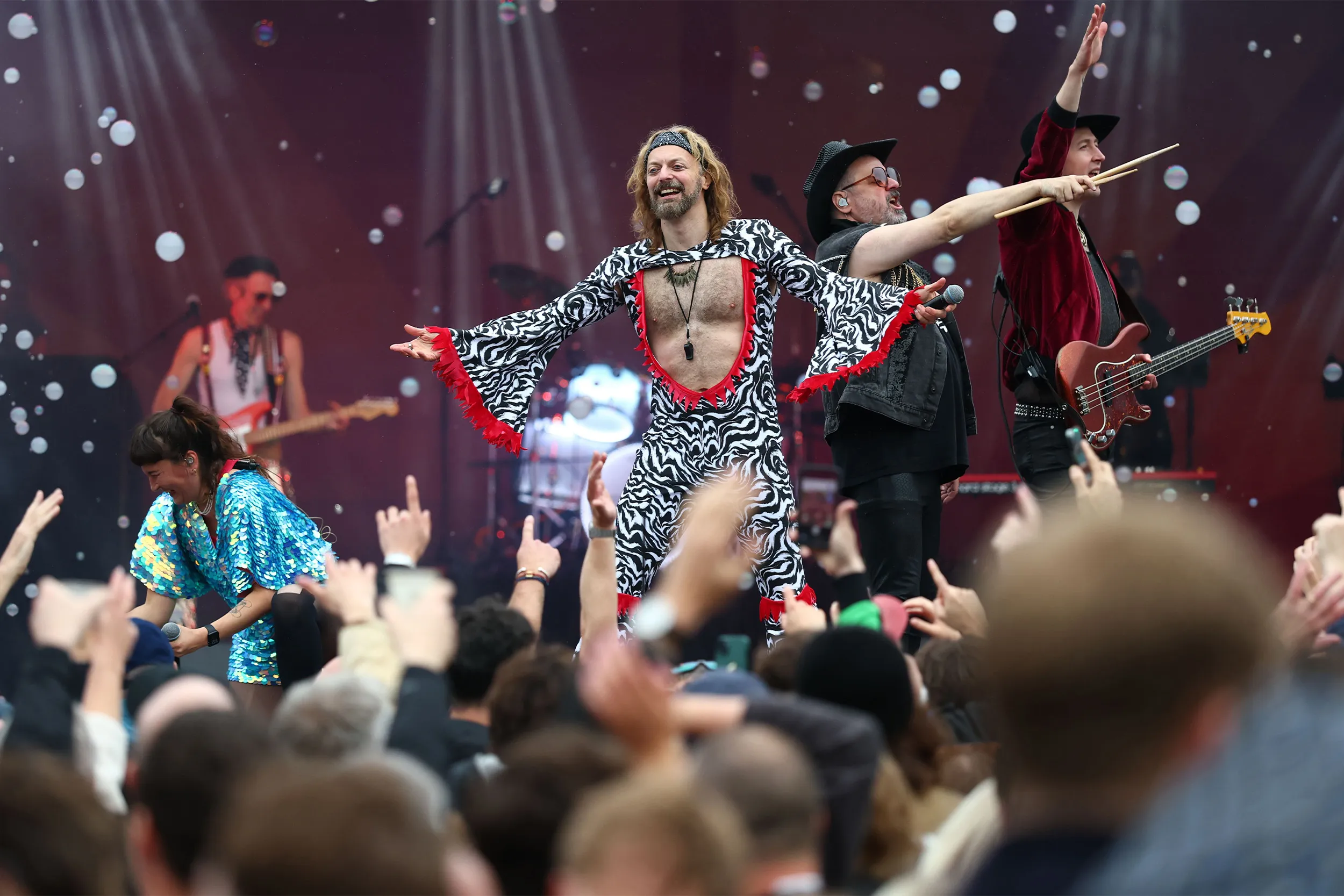 A frontman in a zebra-print flared jumpsuit pointing out over the crowd, bubbles drifting across the stage