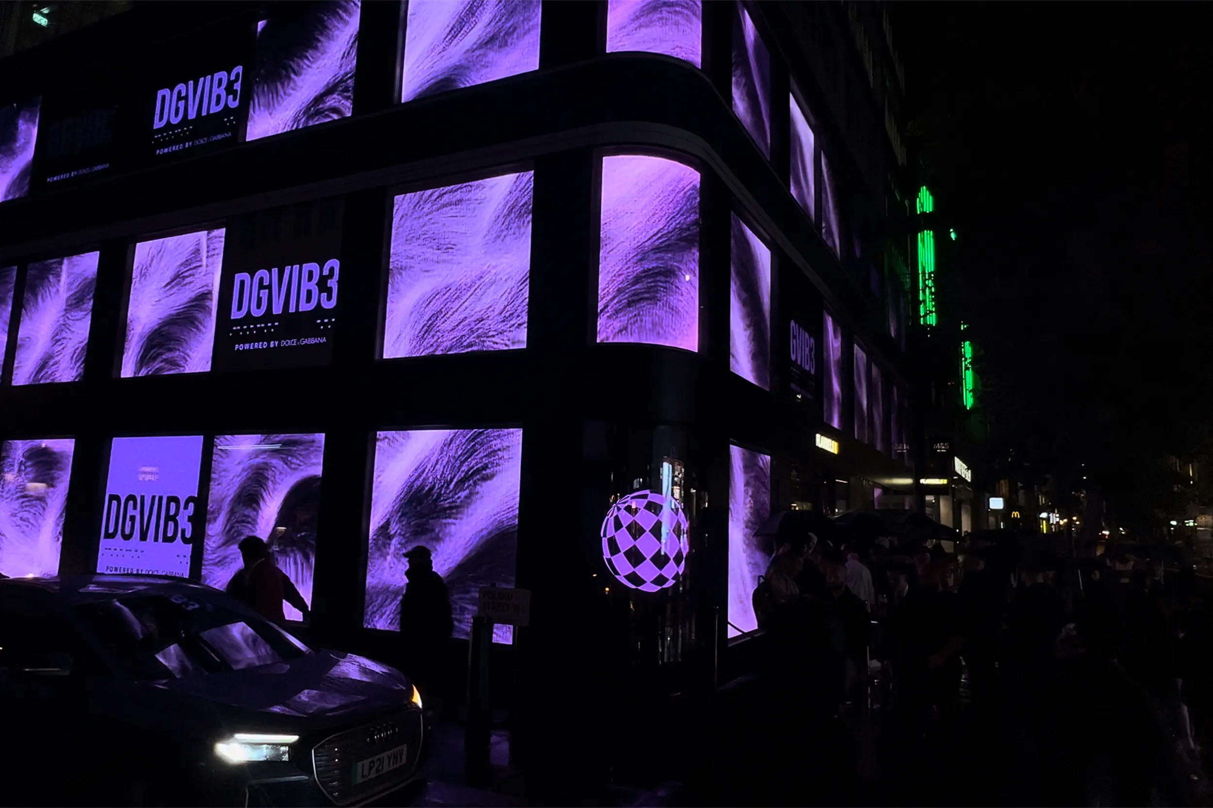 A curved building wrapped in screens of purple feather visuals, a car passing in the dark street
