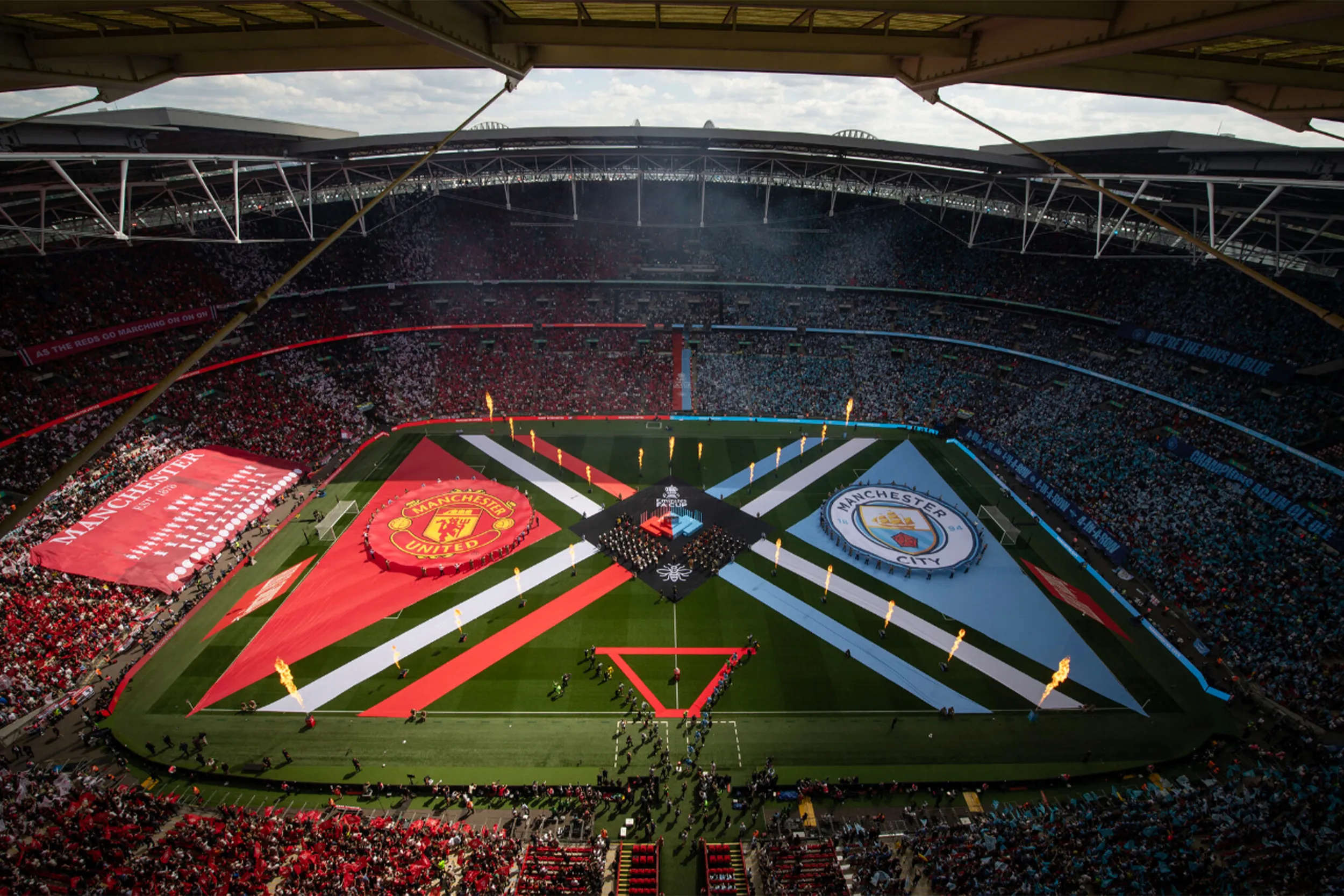 Looking down on the pitch display from high in the stadium: two club crests on red and sky blue, crossed with white bands