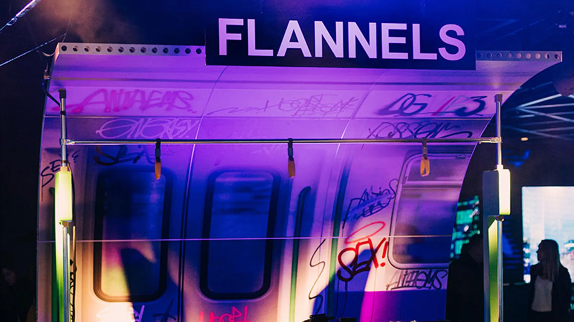 A neon-lit branded structure at a Flannels event, graffitied panels glowing purple