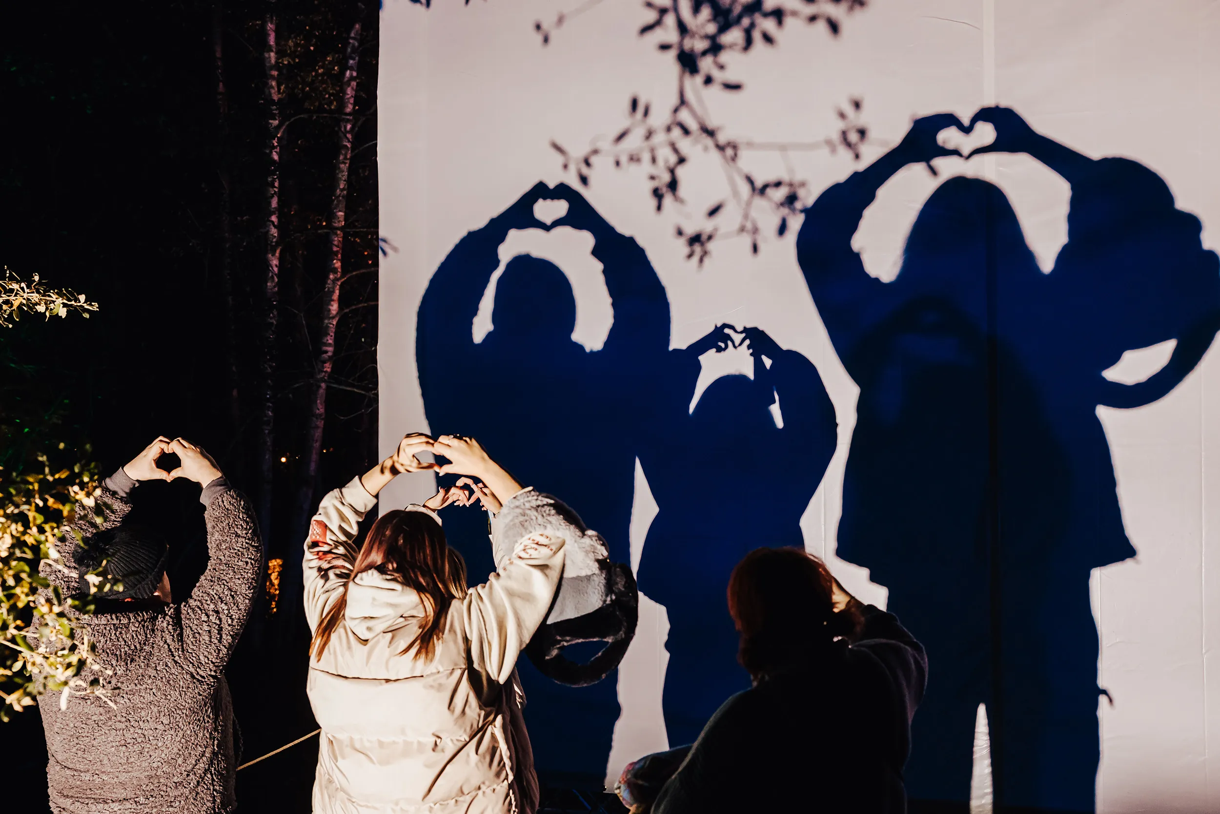 Visitors cast heart-shaped shadows with their hands against an illuminated screen at night