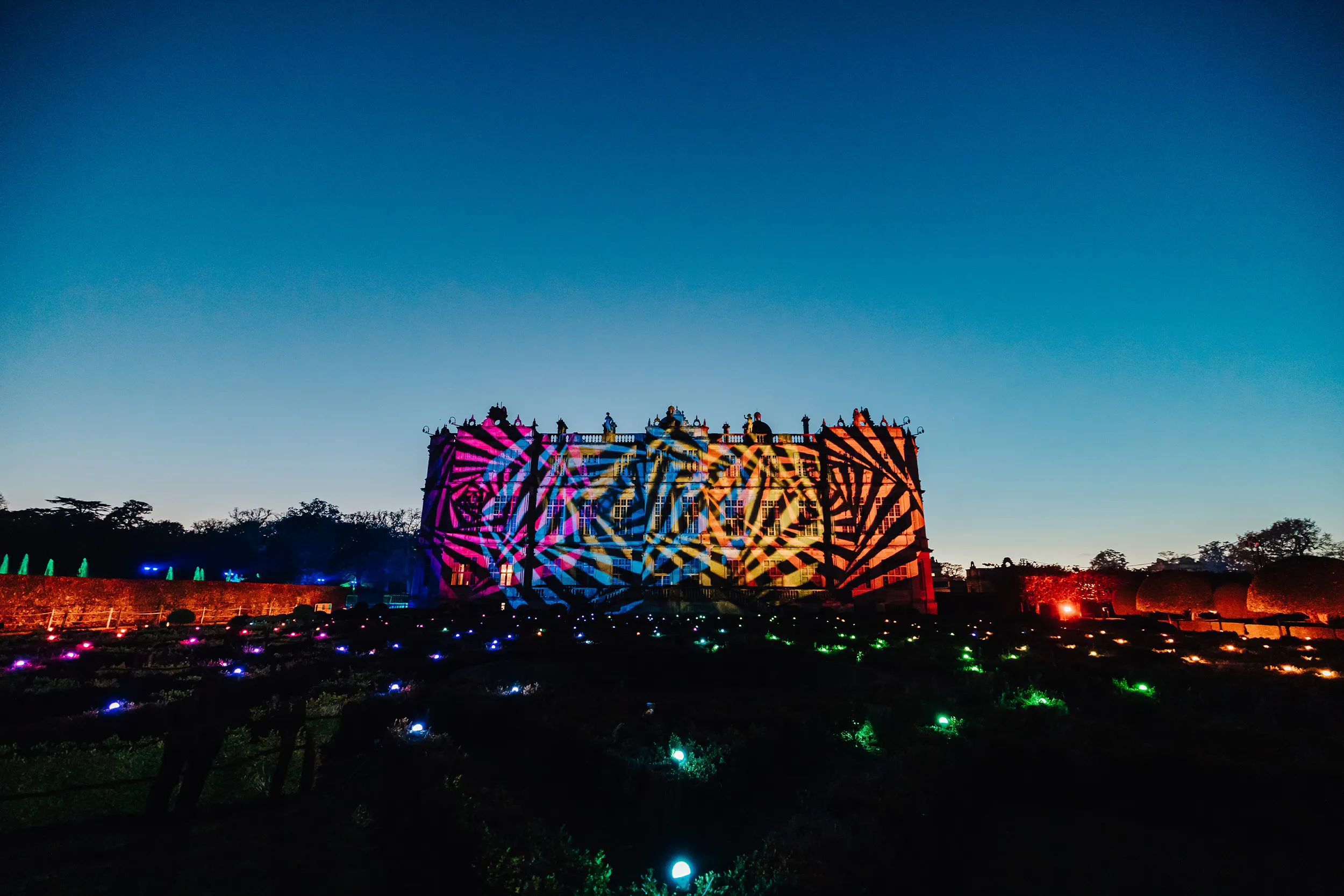 A building across the grounds projected with bold colourful patterns at dusk, small lights in the garden