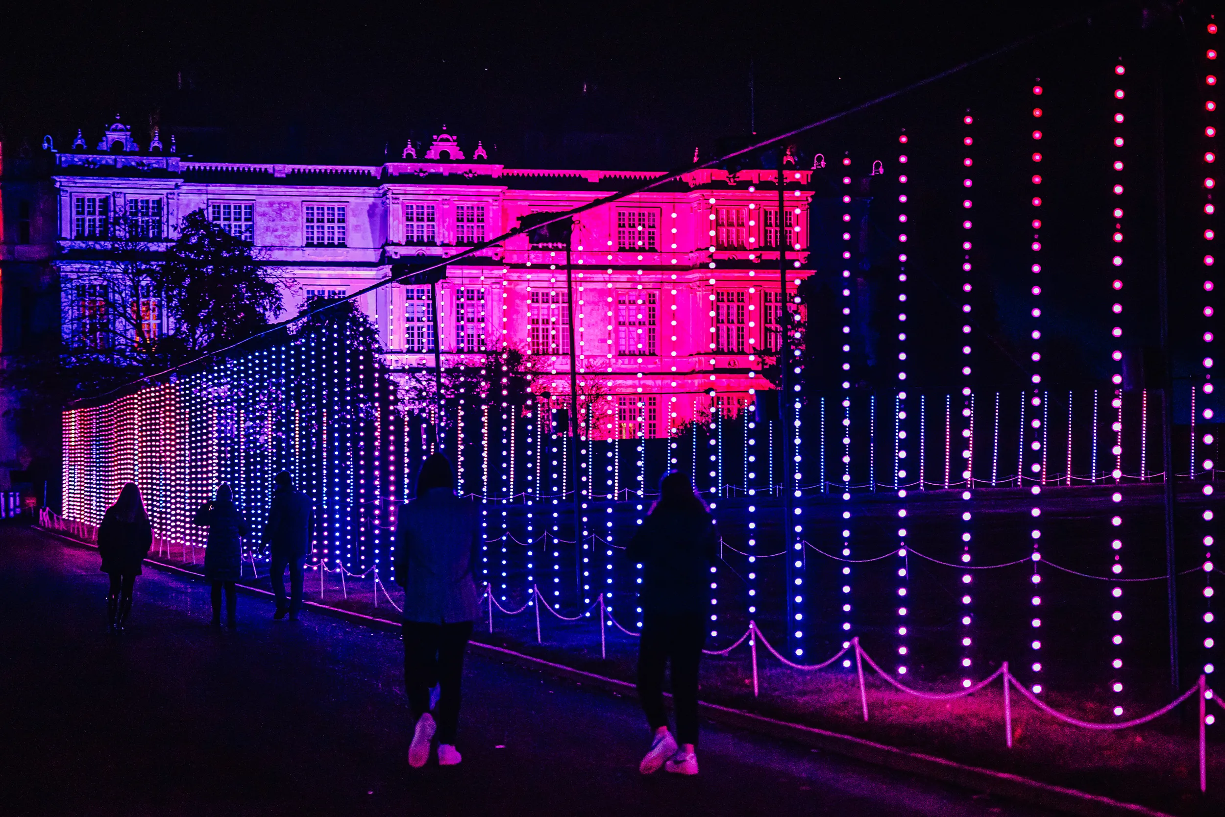 Longleat House lit pink and violet behind a wall of hanging light strings, visitors walking past