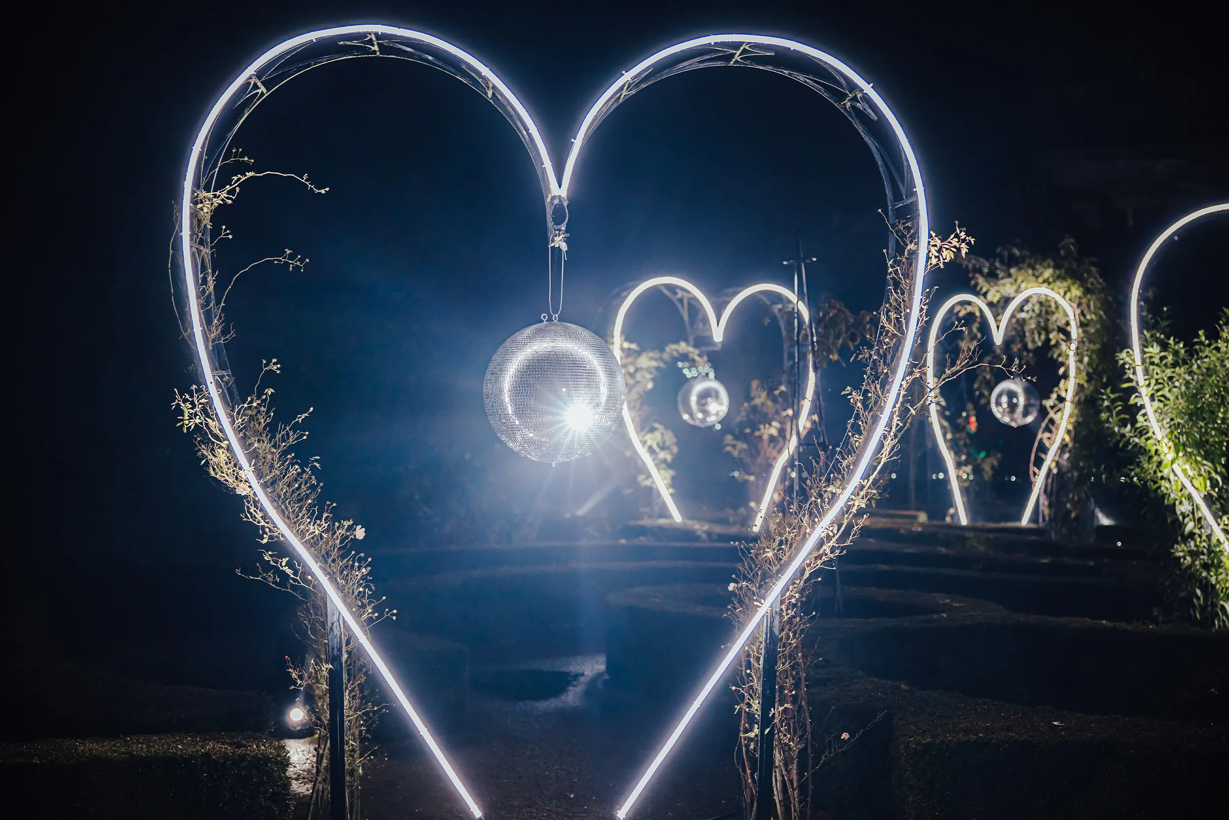 White neon heart arches over a garden path, a mirror ball glinting inside