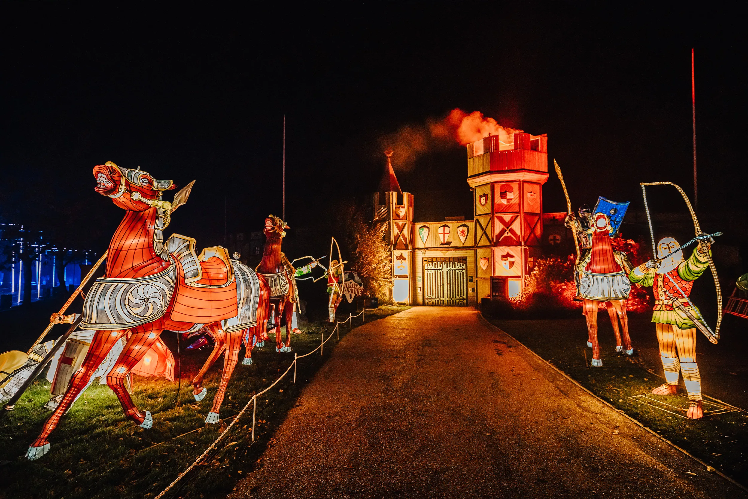Lantern knights on horseback and archers either side of a path leading to a lit castle gate