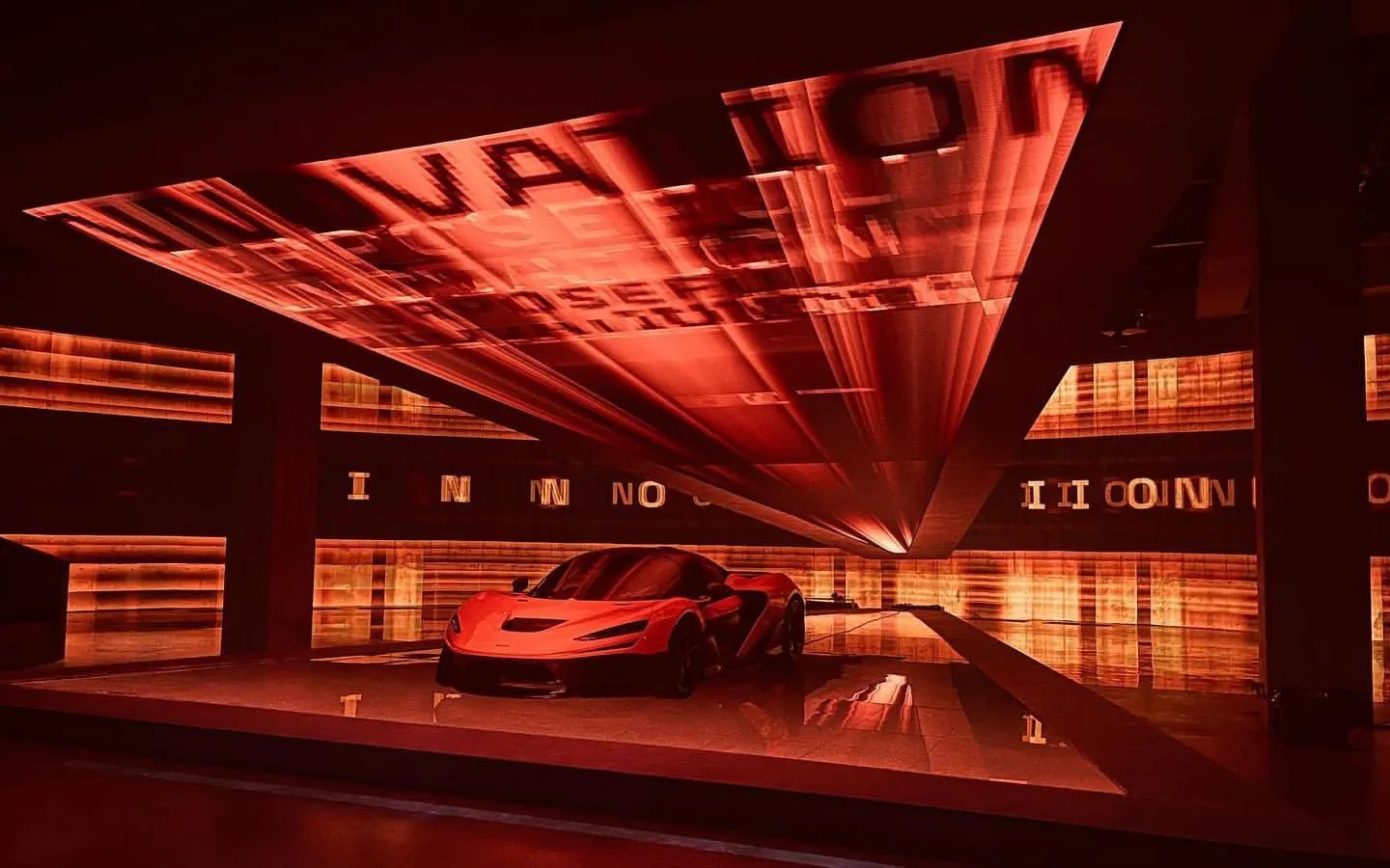 A red McLaren on display beneath a large angled screen in an immersive red-lit installation