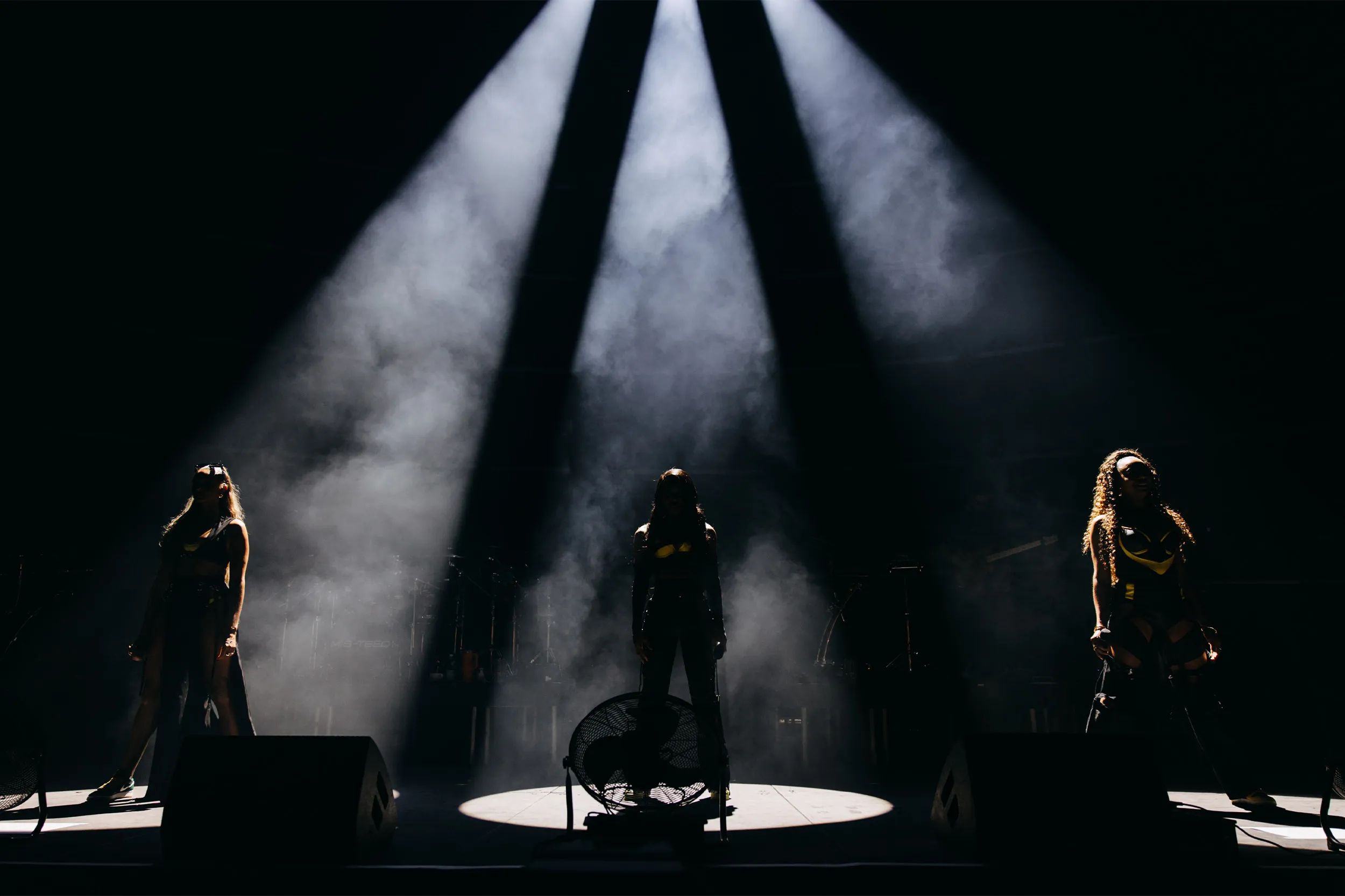 The three members of Mis-Teeq in silhouette, each under a hard white beam cutting through haze