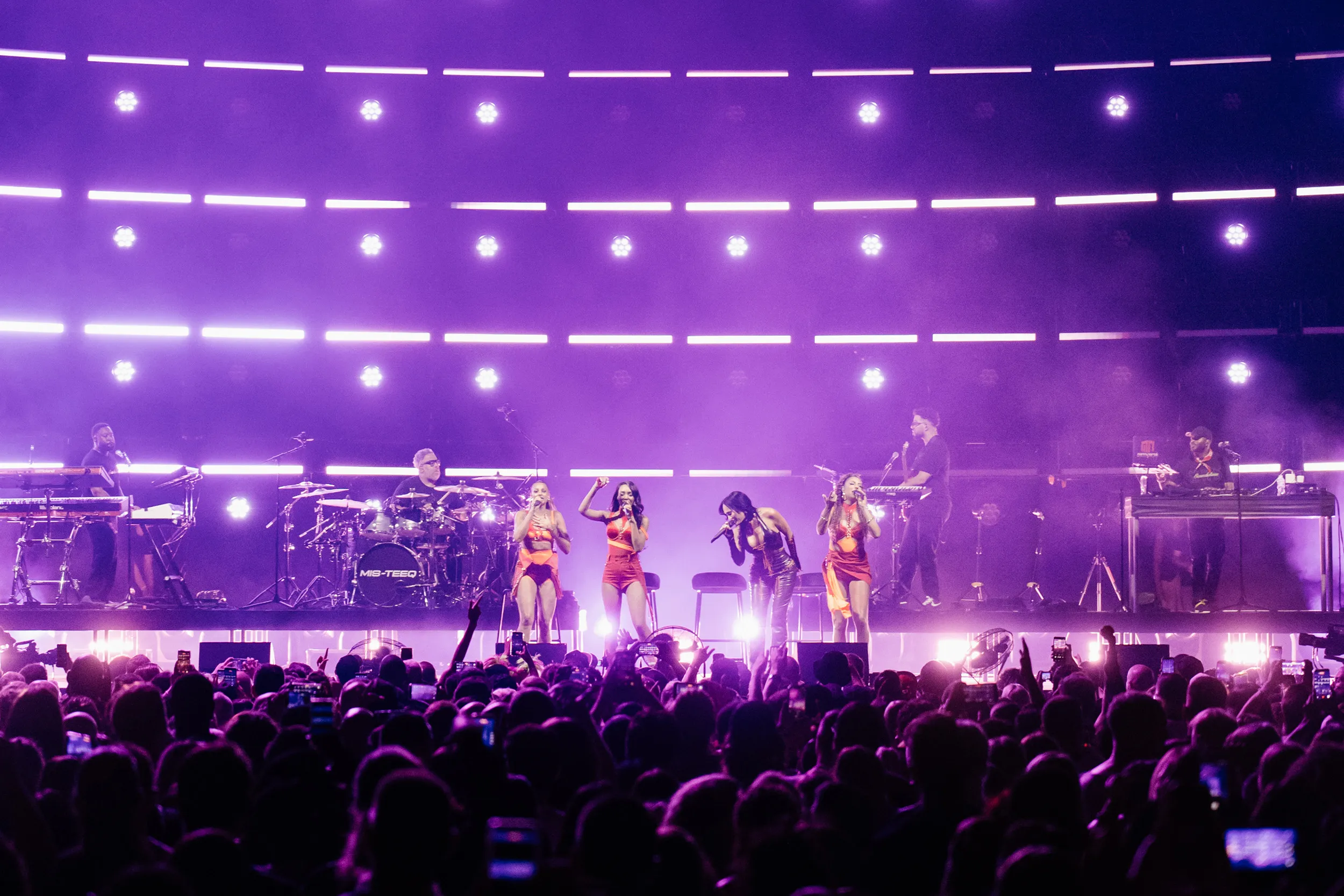 The group and band across the stage under purple light, a row of white points overhead