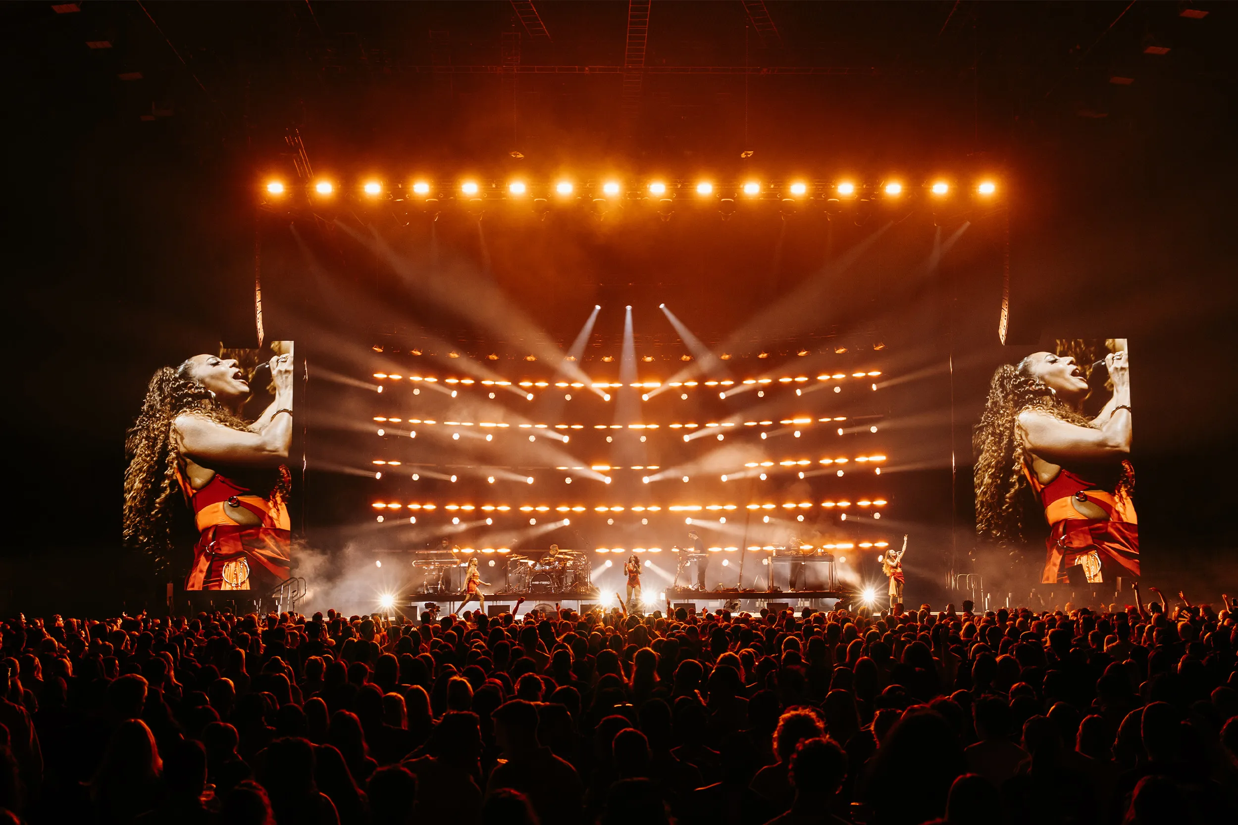 A singer in close-up on both side screens, the stage glowing amber between them