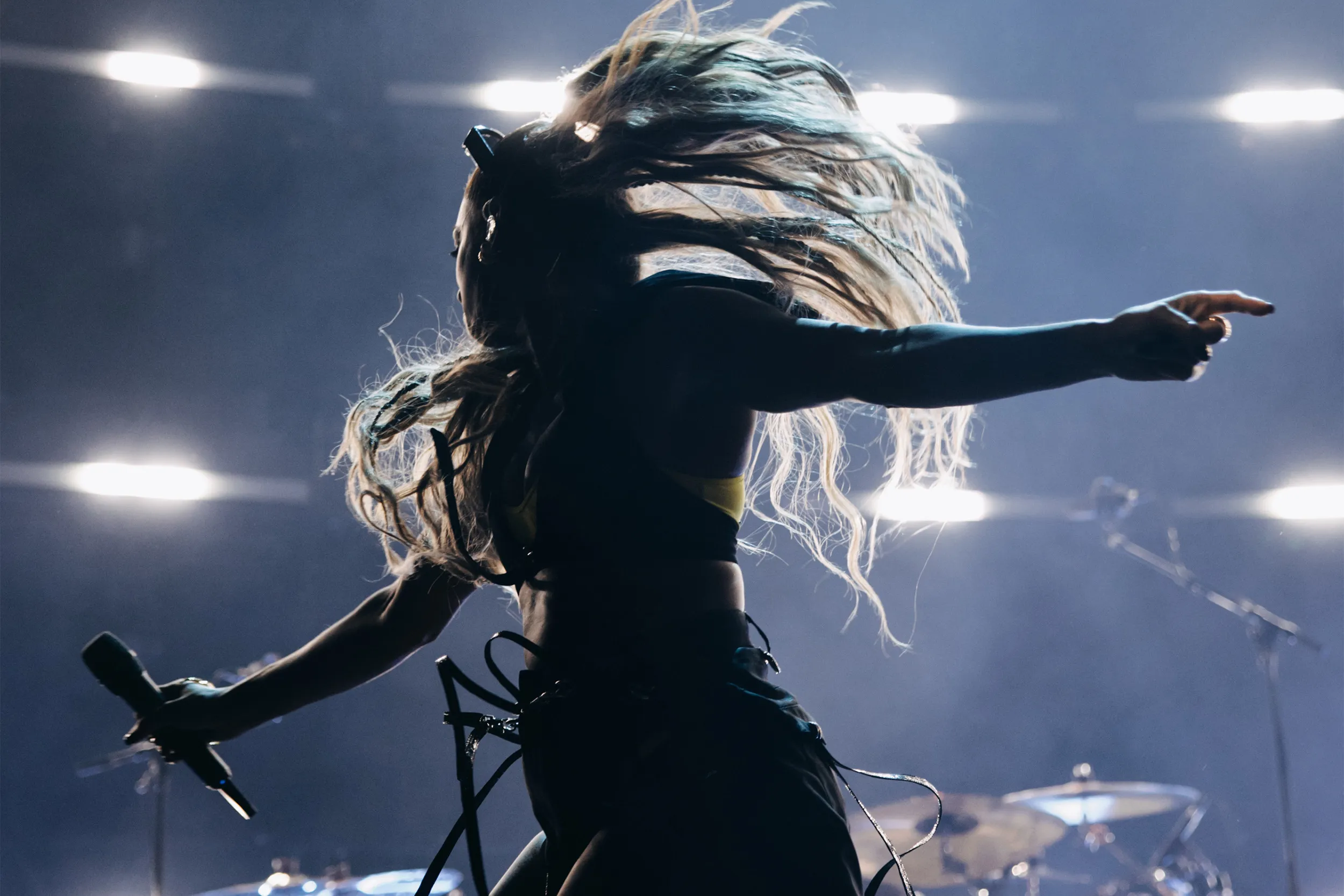 A singer mid hair flip in silhouette, one arm pointing out, rows of white light behind the drum kit