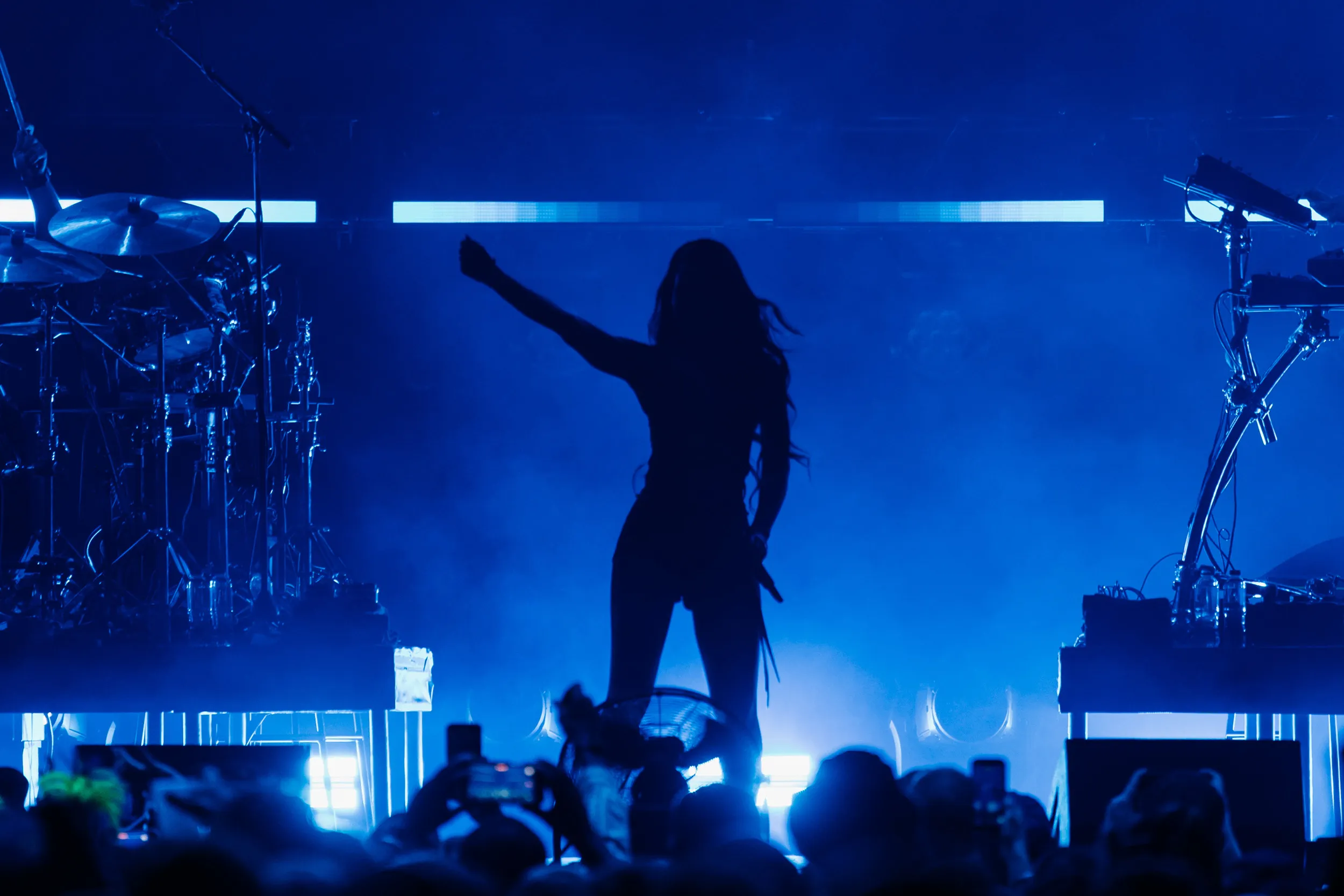 A singer silhouetted with one arm raised, the stage washed in deep blue, phones up in the front row
