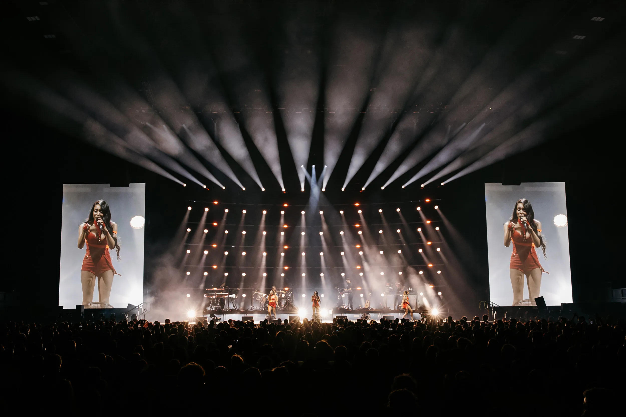 A fan of white beams opening above the stage, a singer in red on the screens either side