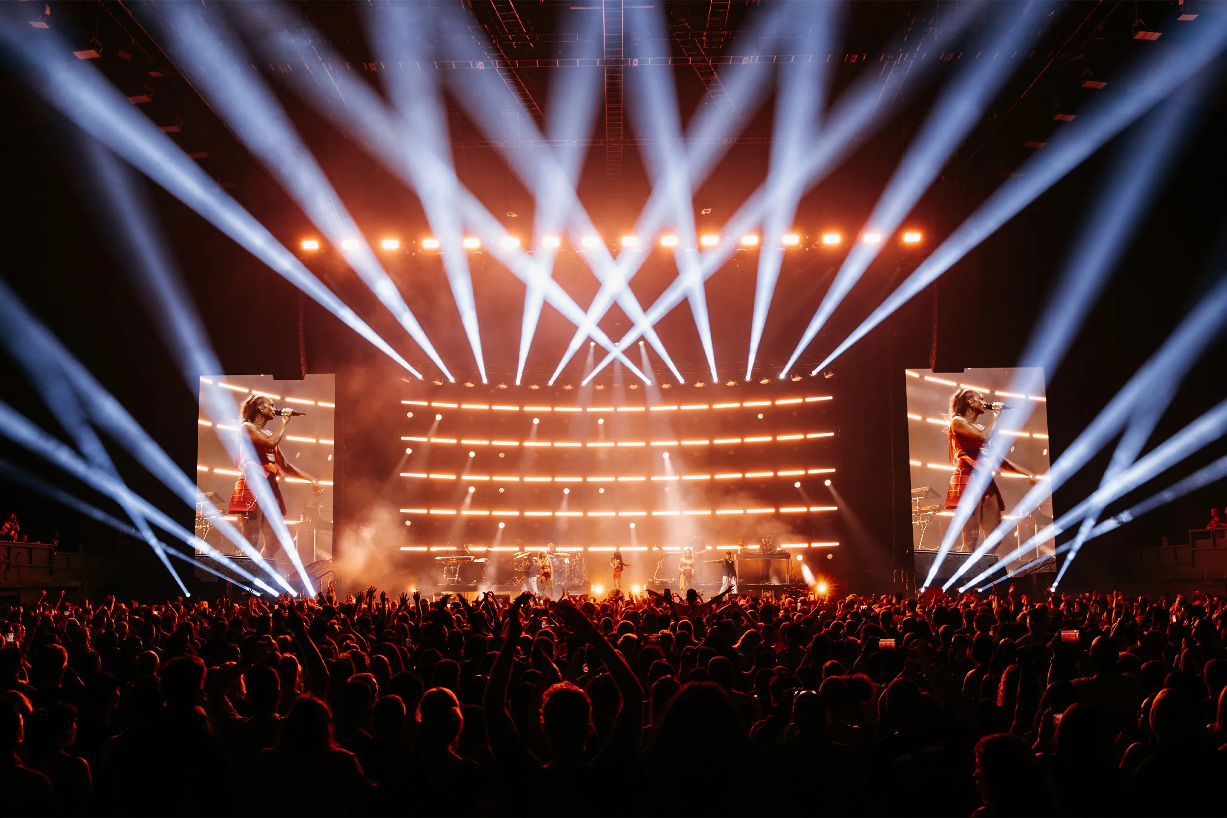 Criss-crossing white beams over an amber stage, the singers on the side screens and a packed arena