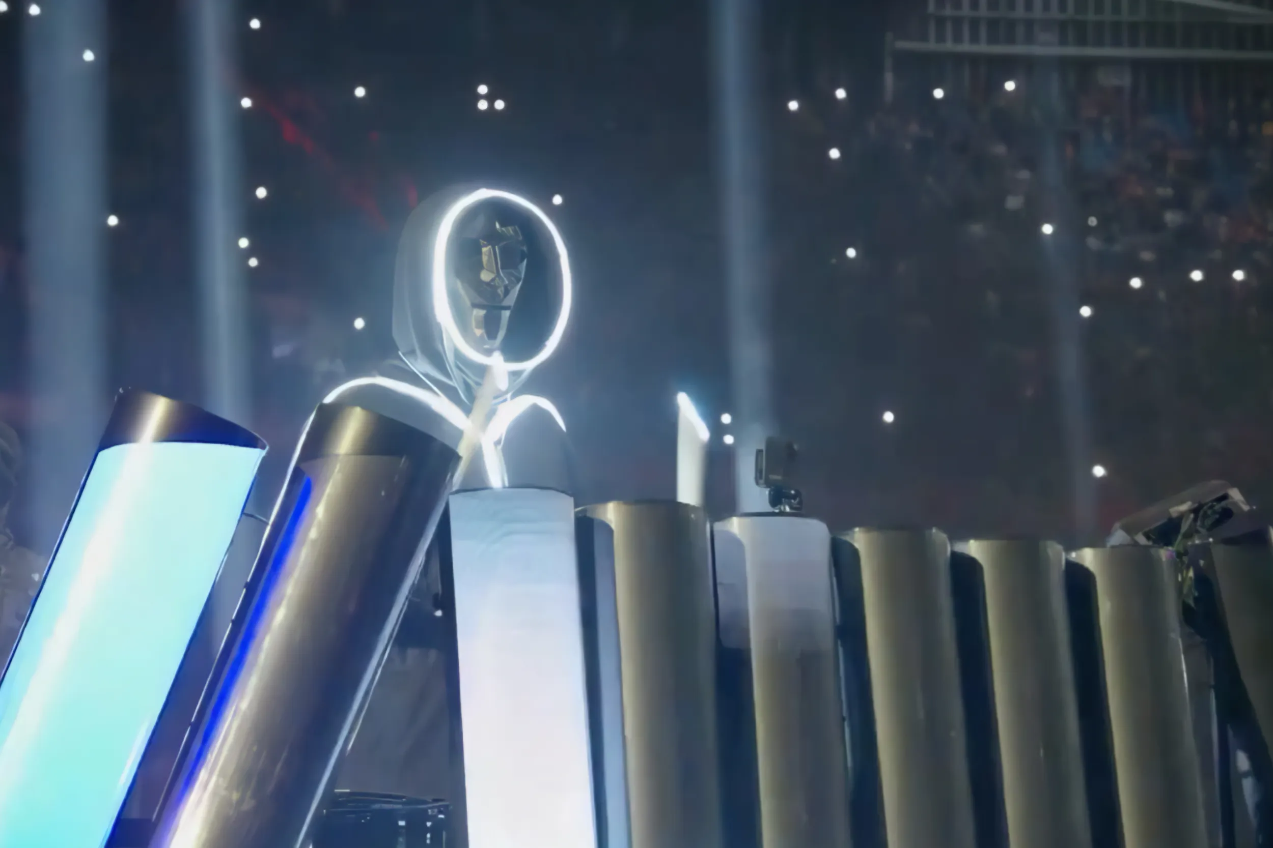 A stickman in a white LED outline behind a row of curved light tubes, phone lights dotting the stands