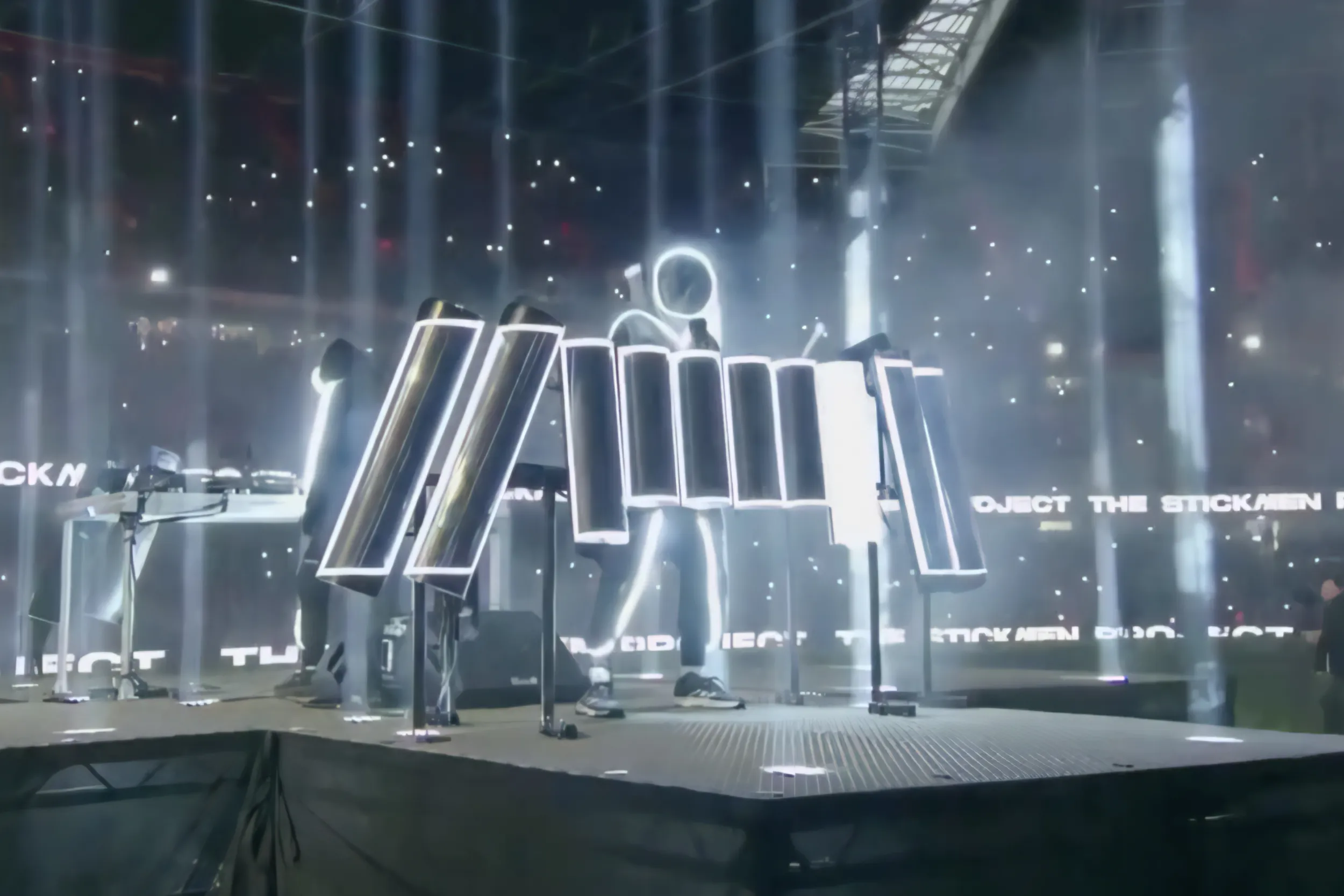The stickmen on stage among angled light panels and white beams, The Stickmen Project scrolling in text behind