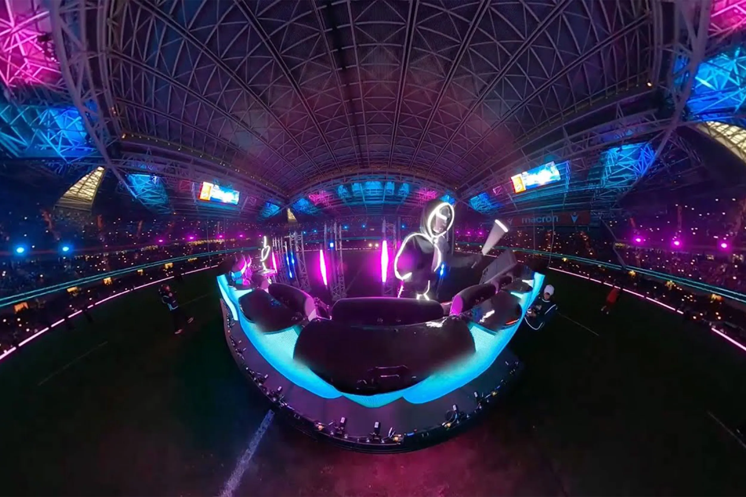 Wide-angle view of the stickmen on a glowing circular stage on the pitch, the stadium roof lit pink and blue above