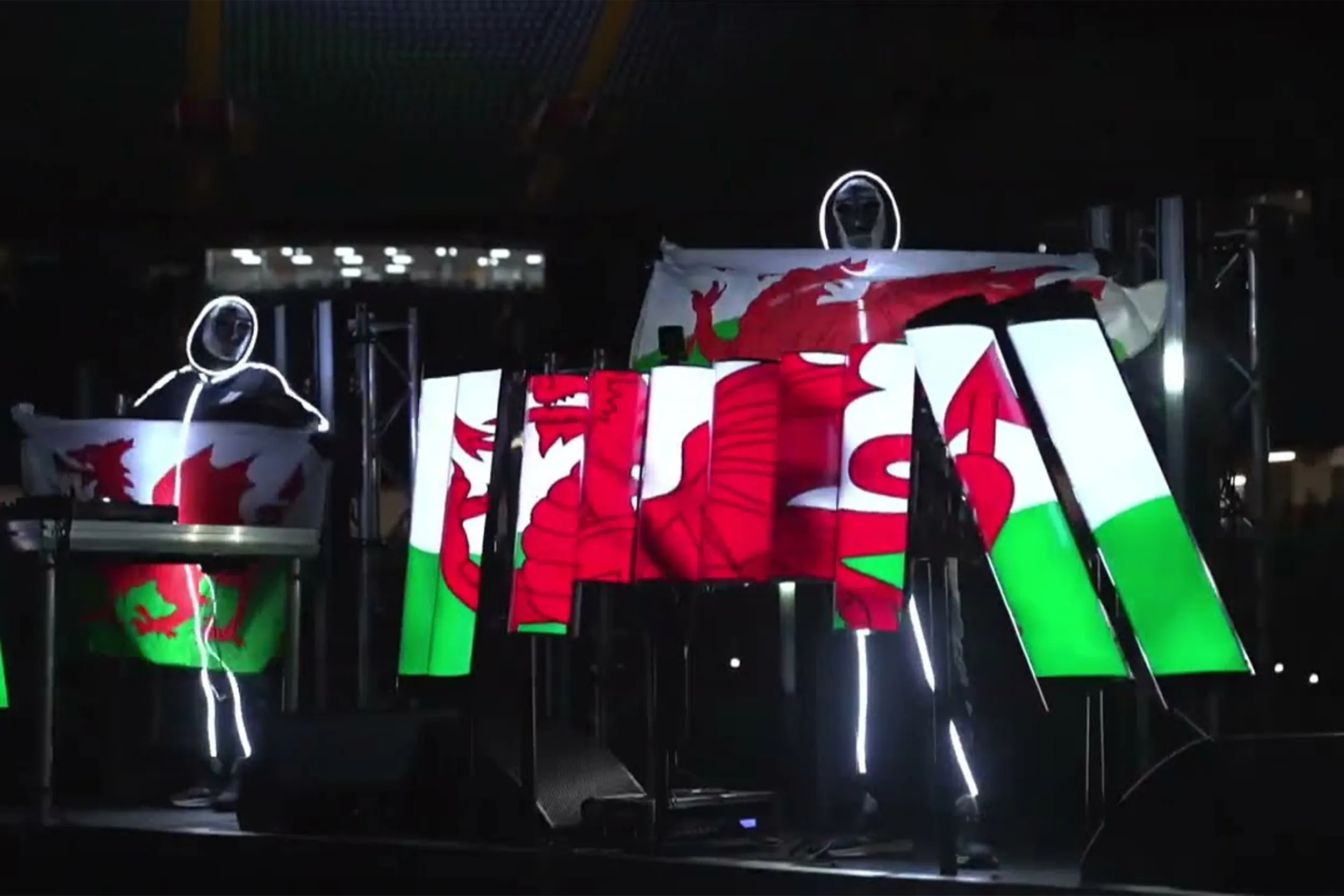 Two illuminated stick-figure performers behind angled light panels showing the Welsh flag, one holding a flag aloft