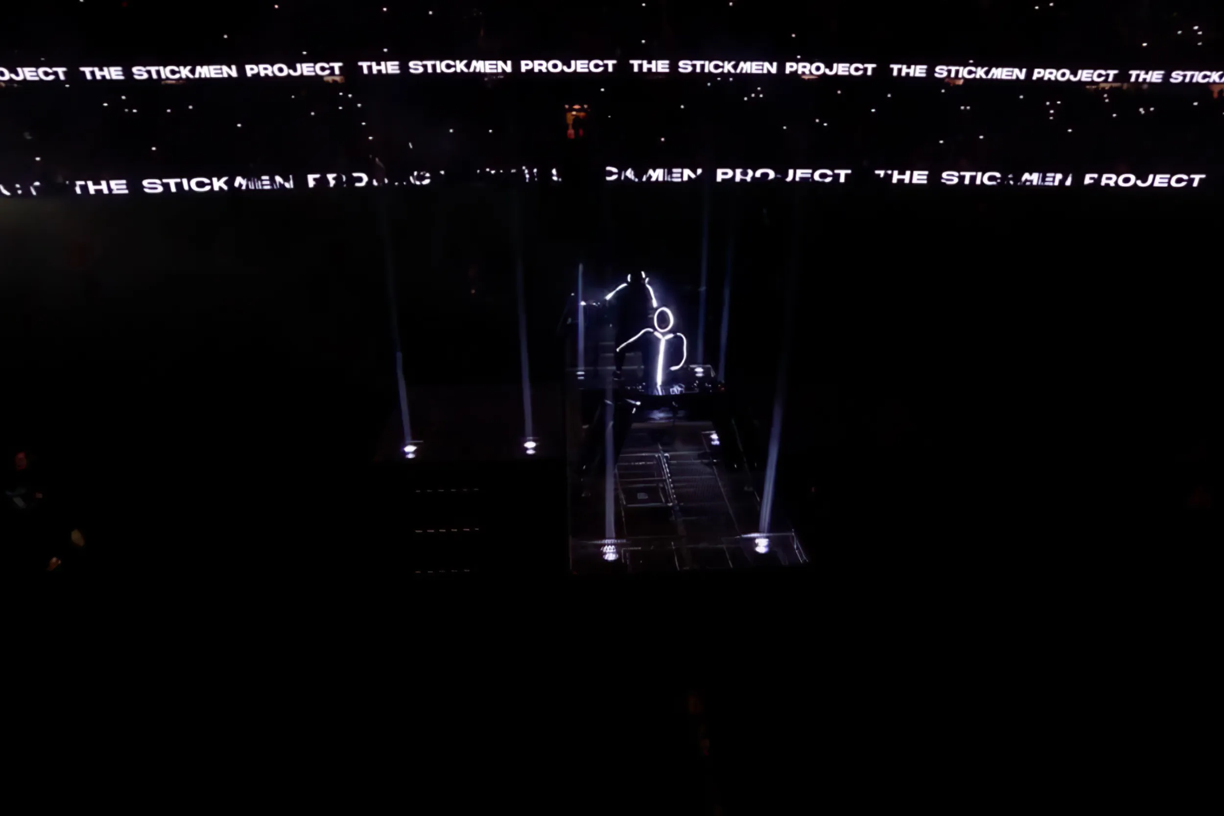 A lone stickman small on the stage in the dark stadium, The Stickmen Project in lines of text above
