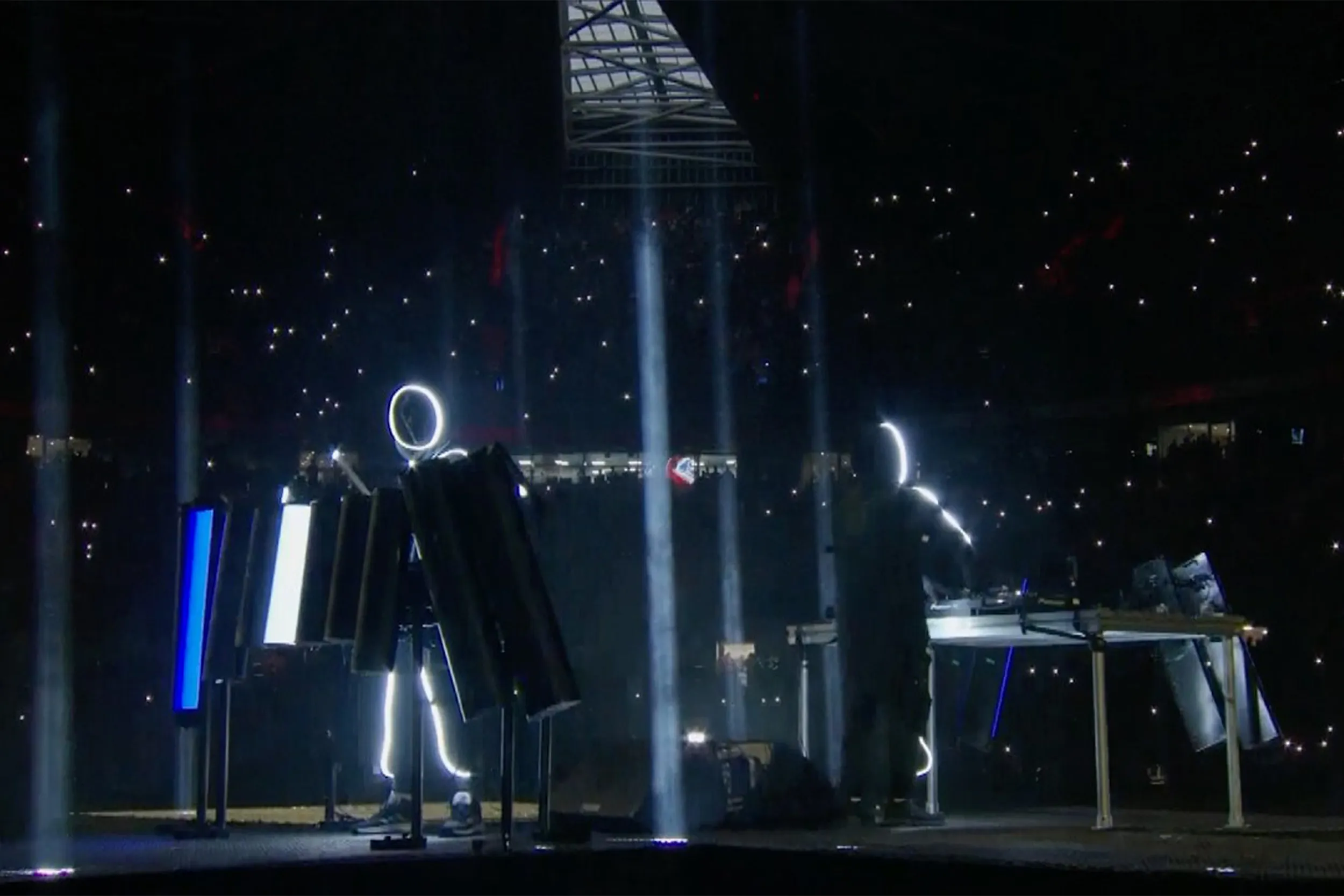 The stickmen performing among upright light panels, white beams rising into the dark stadium