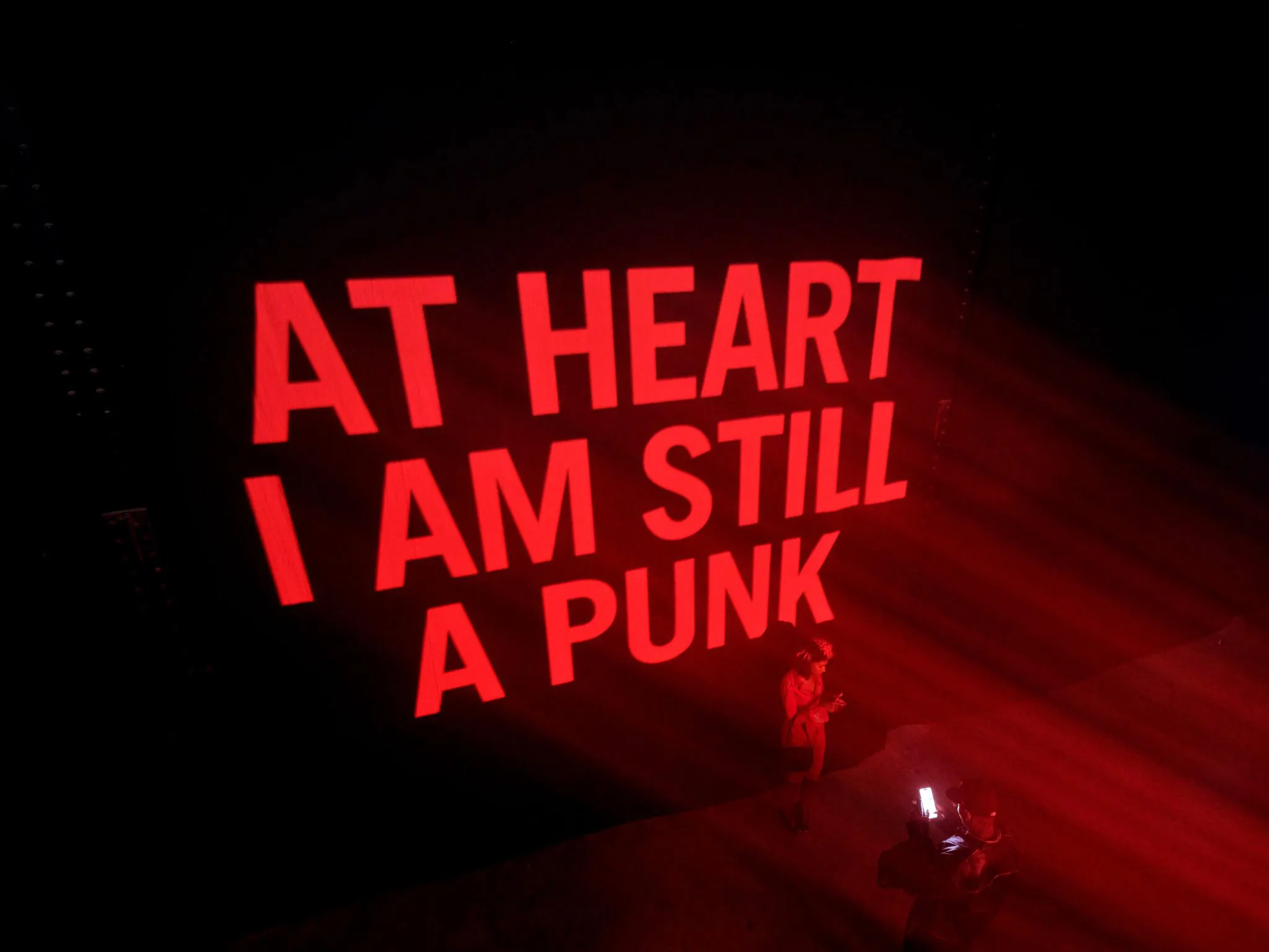 A giant screen reads 'at heart I am still a punk' above the crowd below
