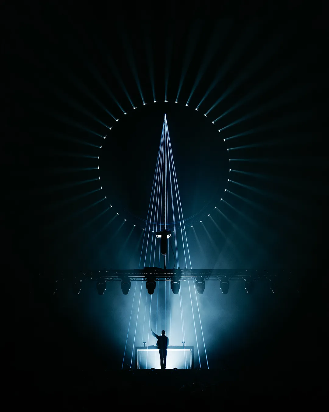Sub Focus alone at the decks beneath a ring of white beams and a single spike of light