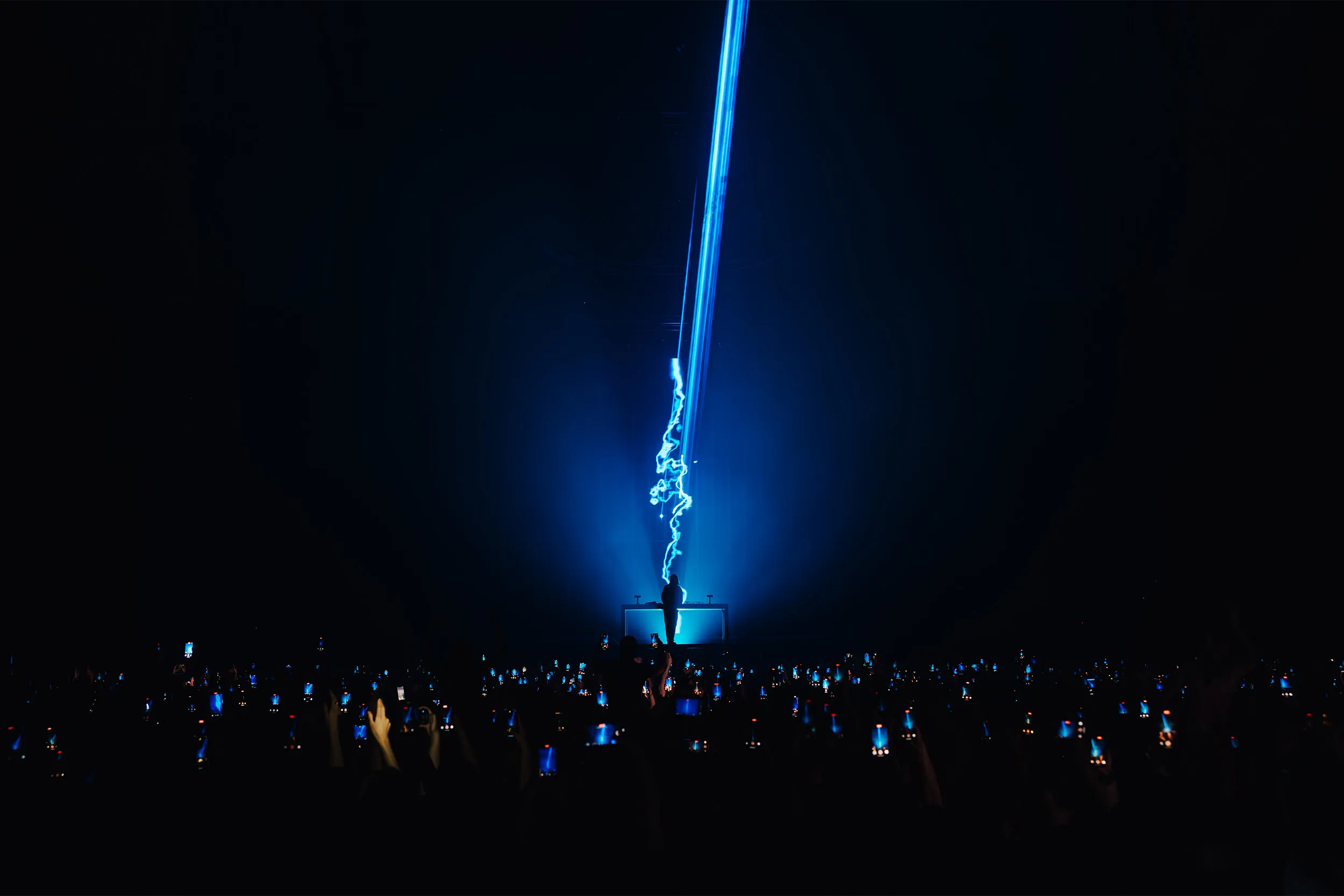 A single blue beam rising from the booth into the dark, the crowd lit by phone screens