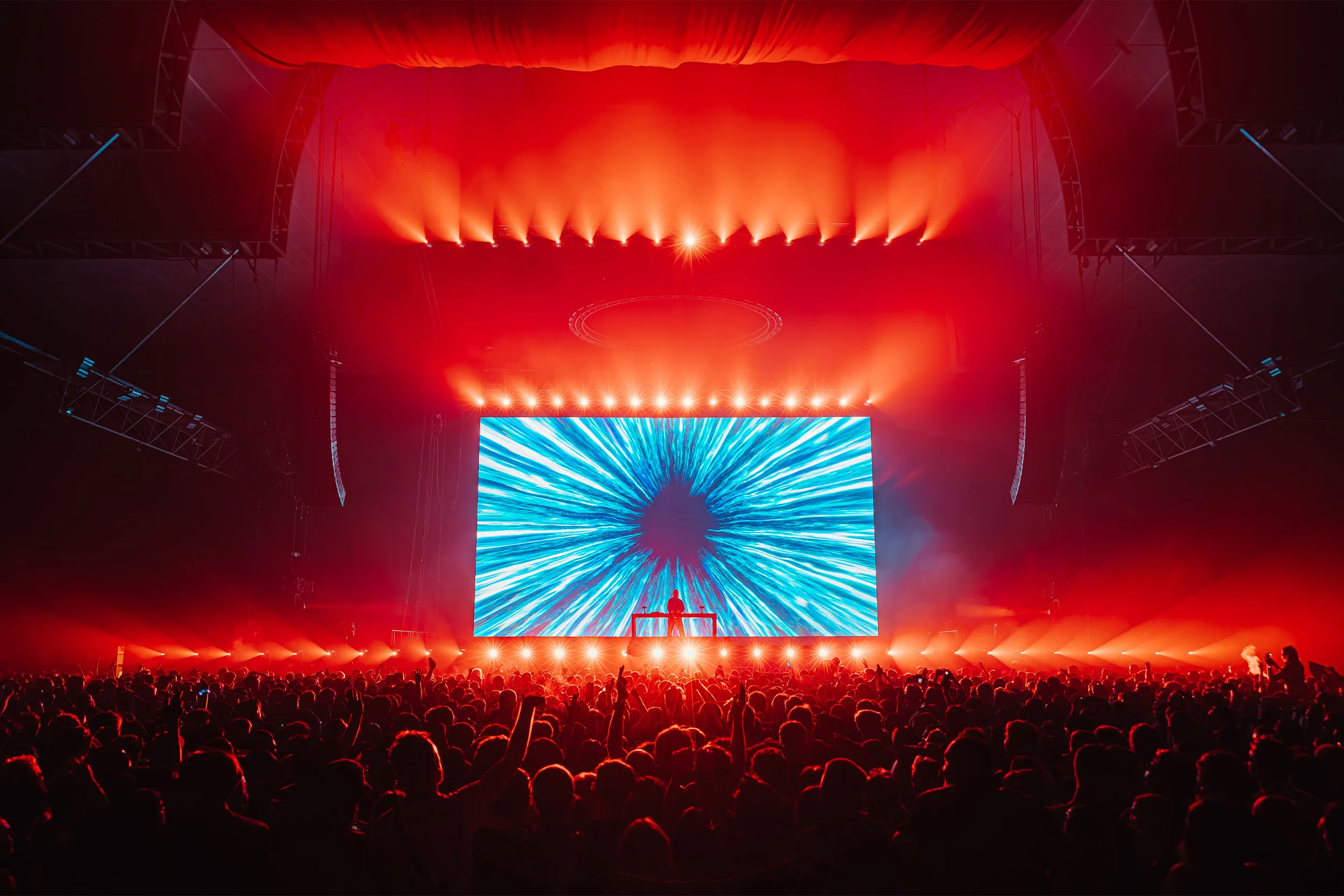 The stage under red light, a blue burst of visuals on the screen behind the booth