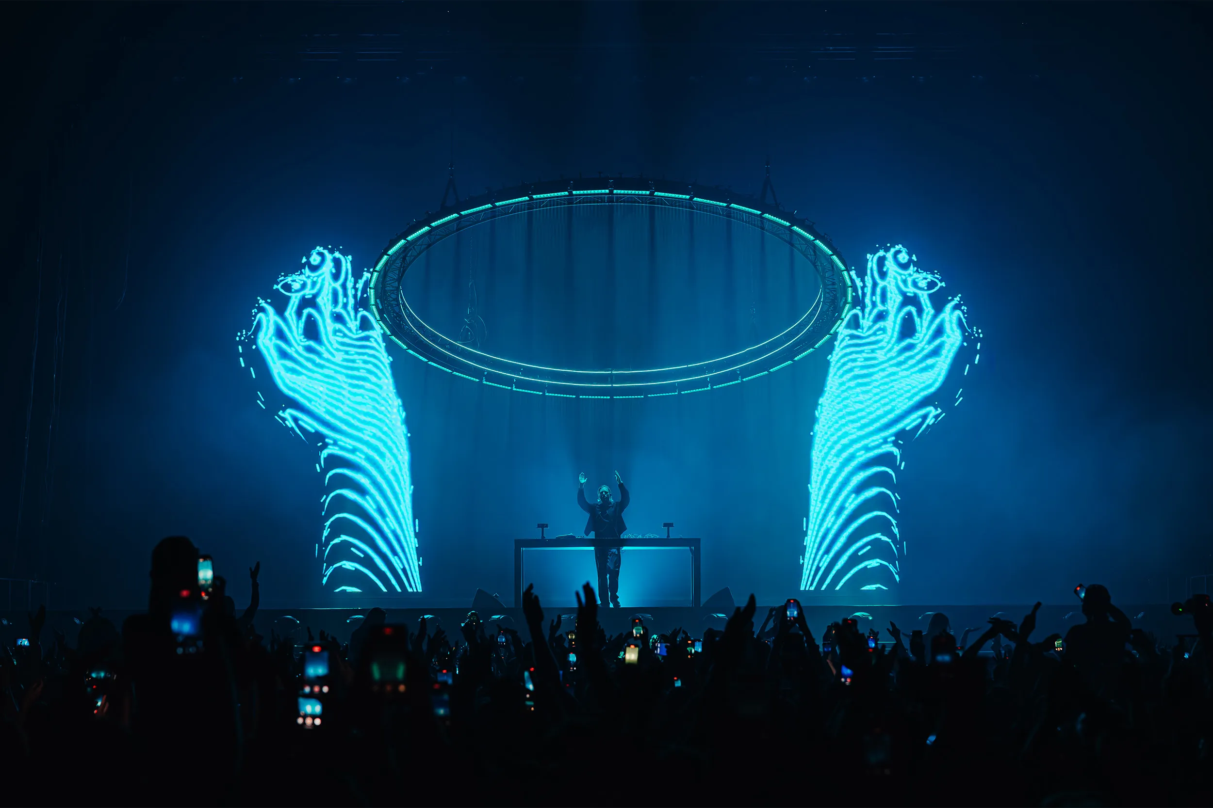Two giant glowing hands in cyan either side of a suspended ring, Sub Focus at the decks between them