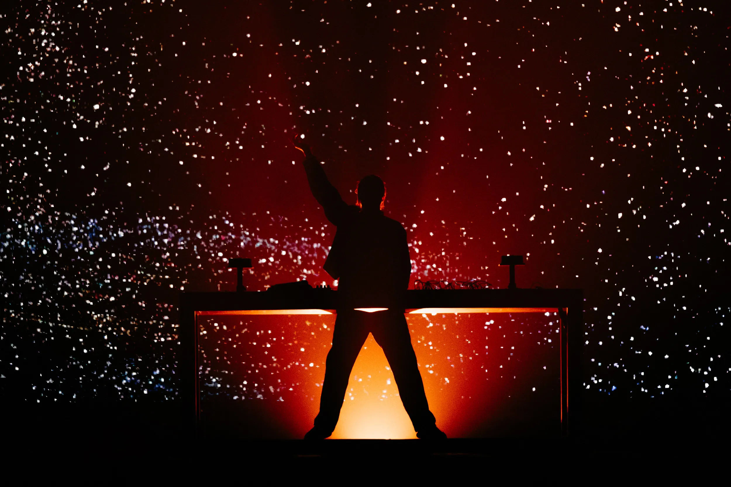 Sub Focus in silhouette at the decks, arm raised, against a field of stars on red
