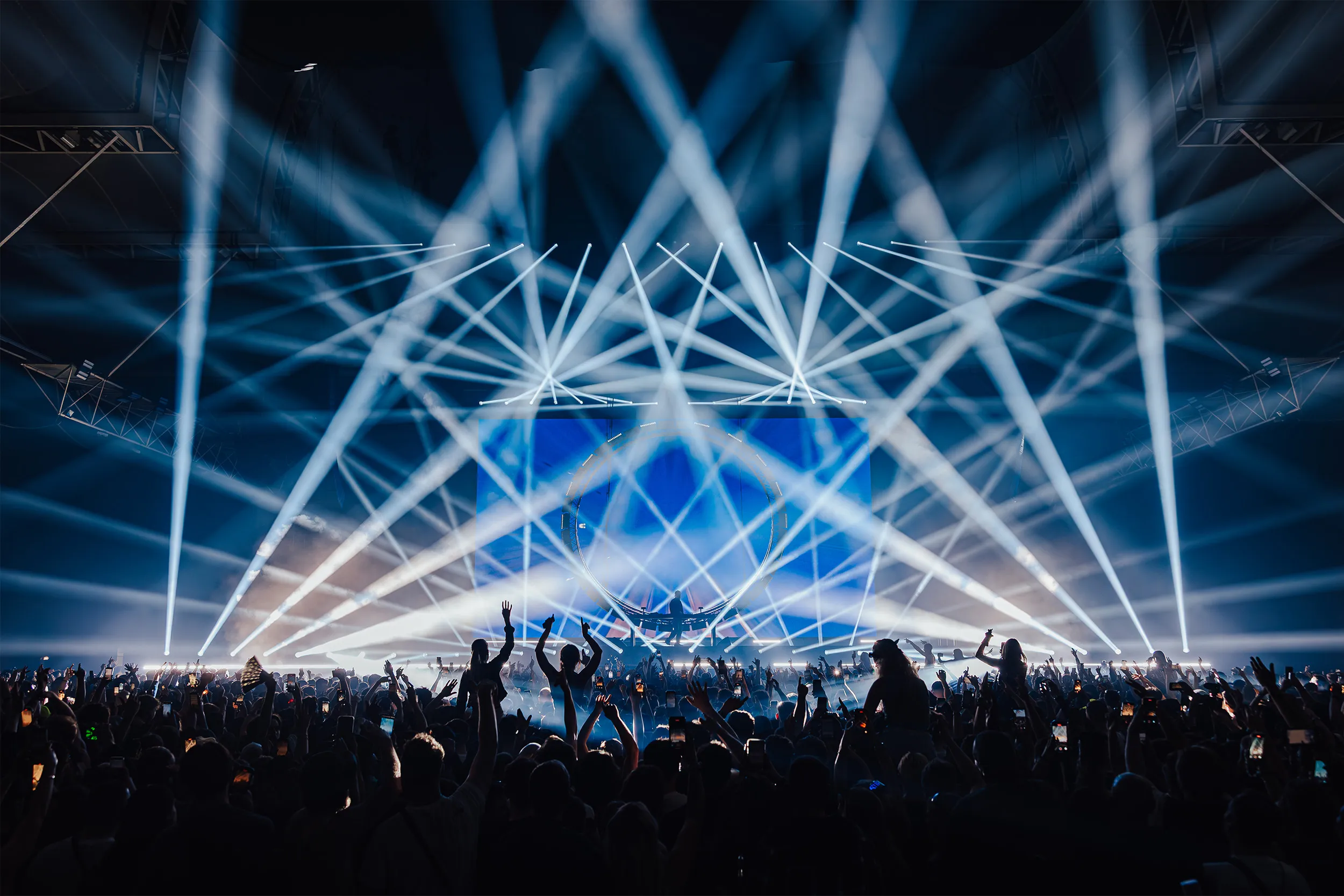 White beams crossing over the stage in every direction above a crowd with hands up