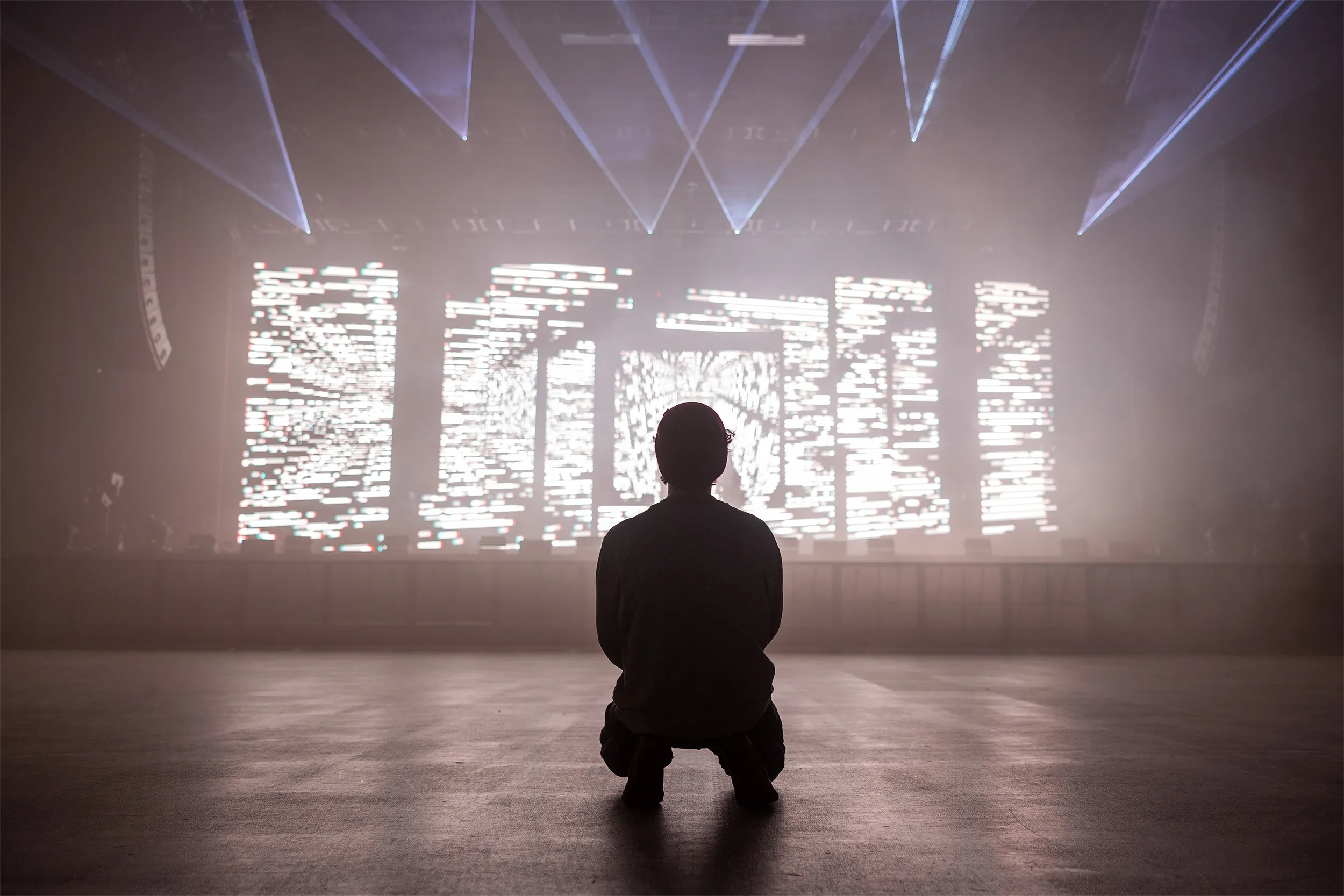 A lone figure kneeling in silhouette before a glitching LED wall, lasers cutting overhead